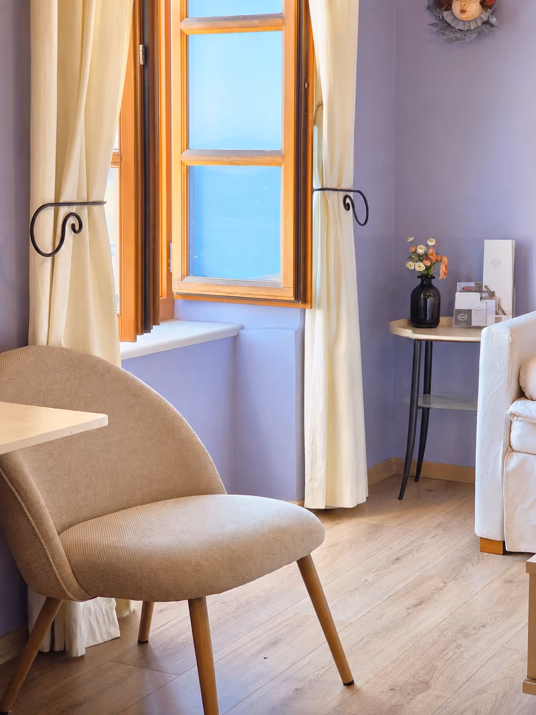 Cozy corner with a beige fabric chair, wooden floor, purple walls, open wooden window, and a small table with flowers.