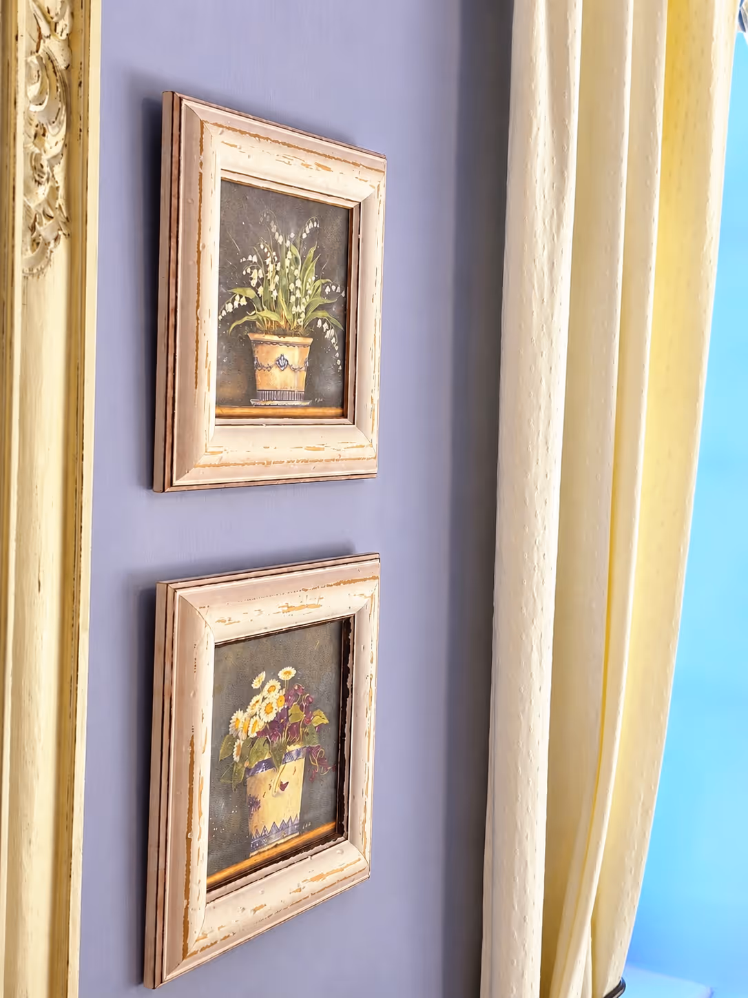 Two framed floral paintings with vintage white frames hanging on a lavender wall near cream curtains.