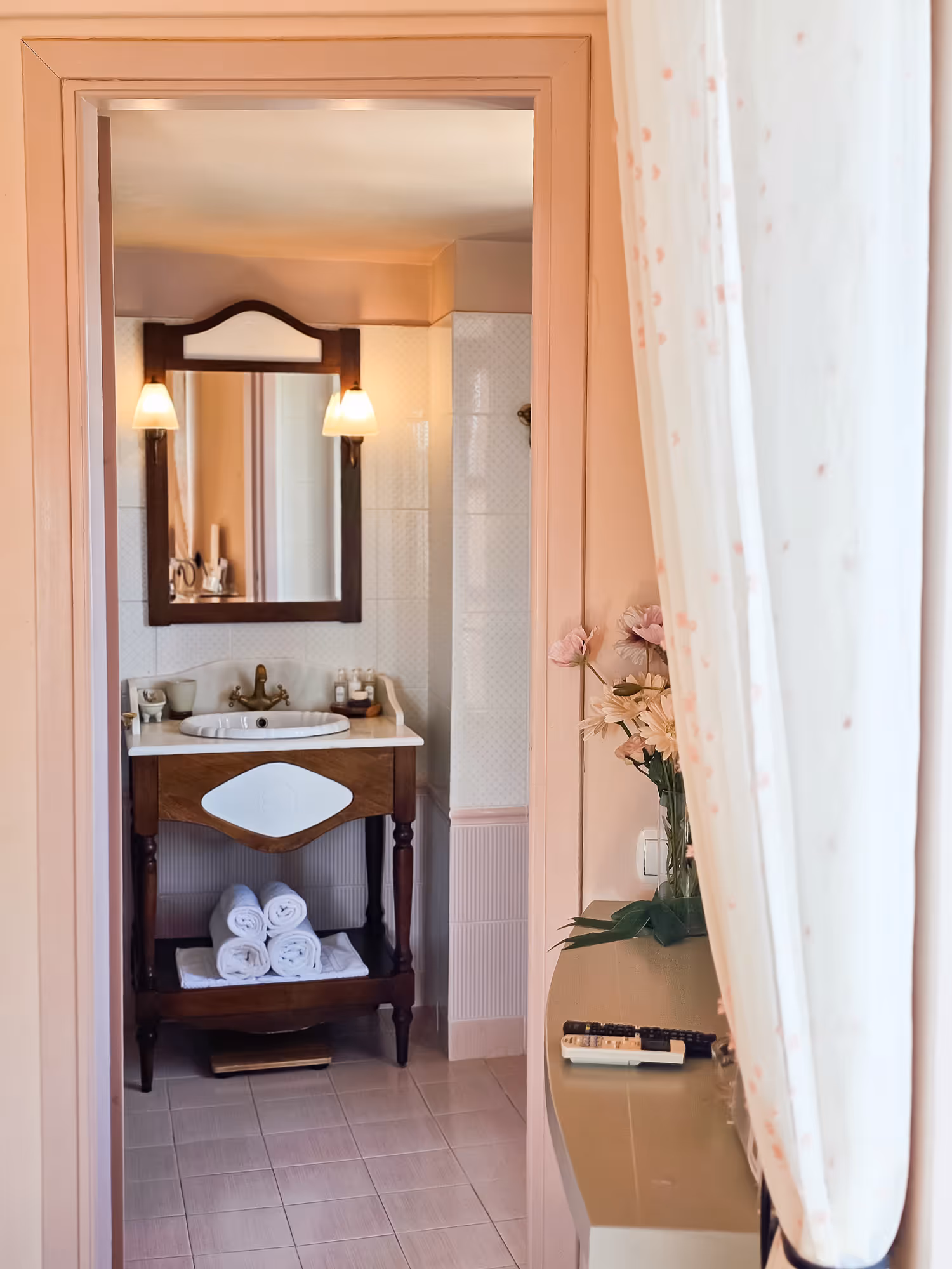 Vintage bathroom sink with mirror, towels below, lit wall sconces, and flowers on a nearby surface.