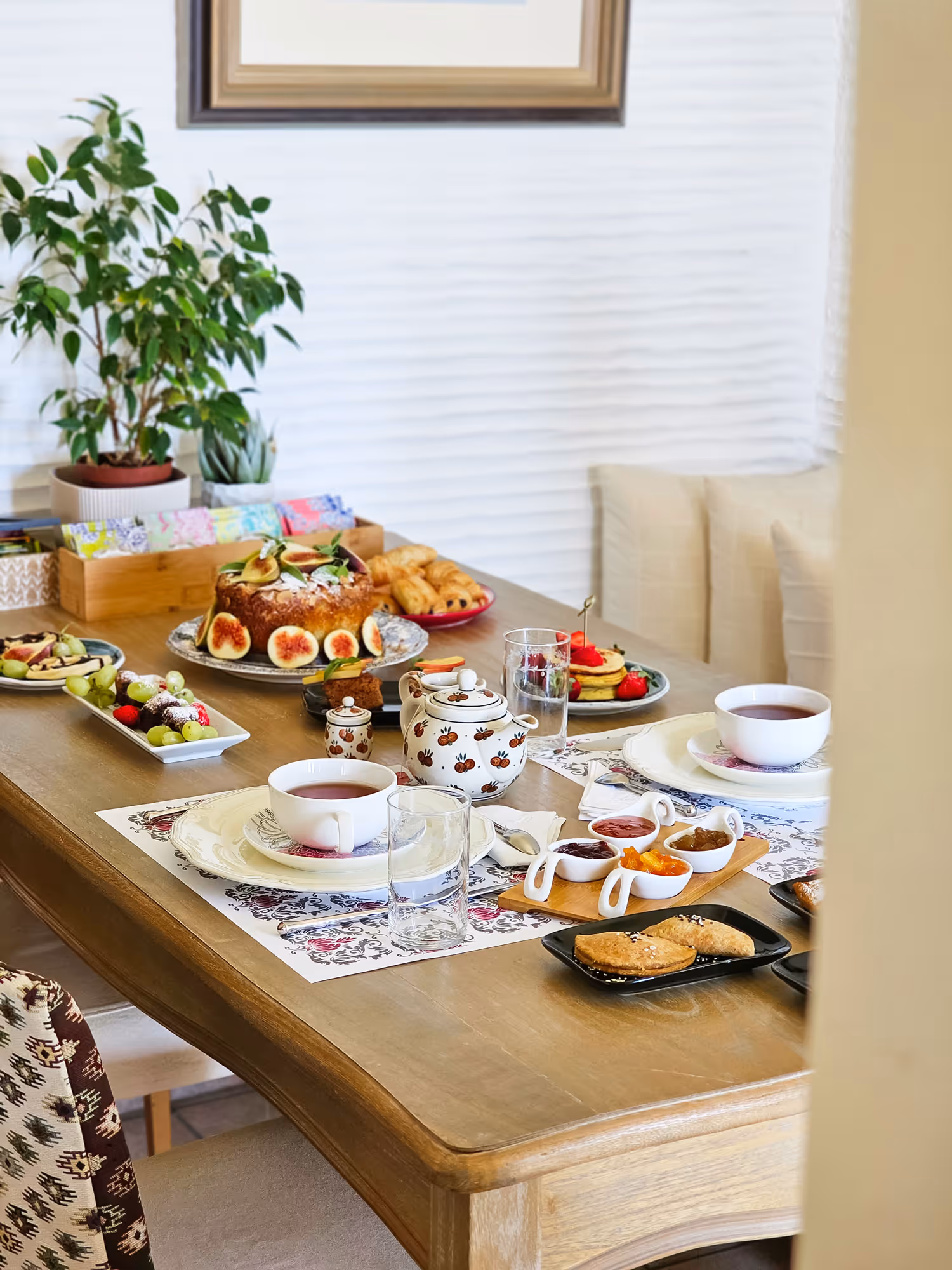 Table set for tea with cups, teapot, cake, pastries, fruit, jams, and water glasses in a cozy dining area.