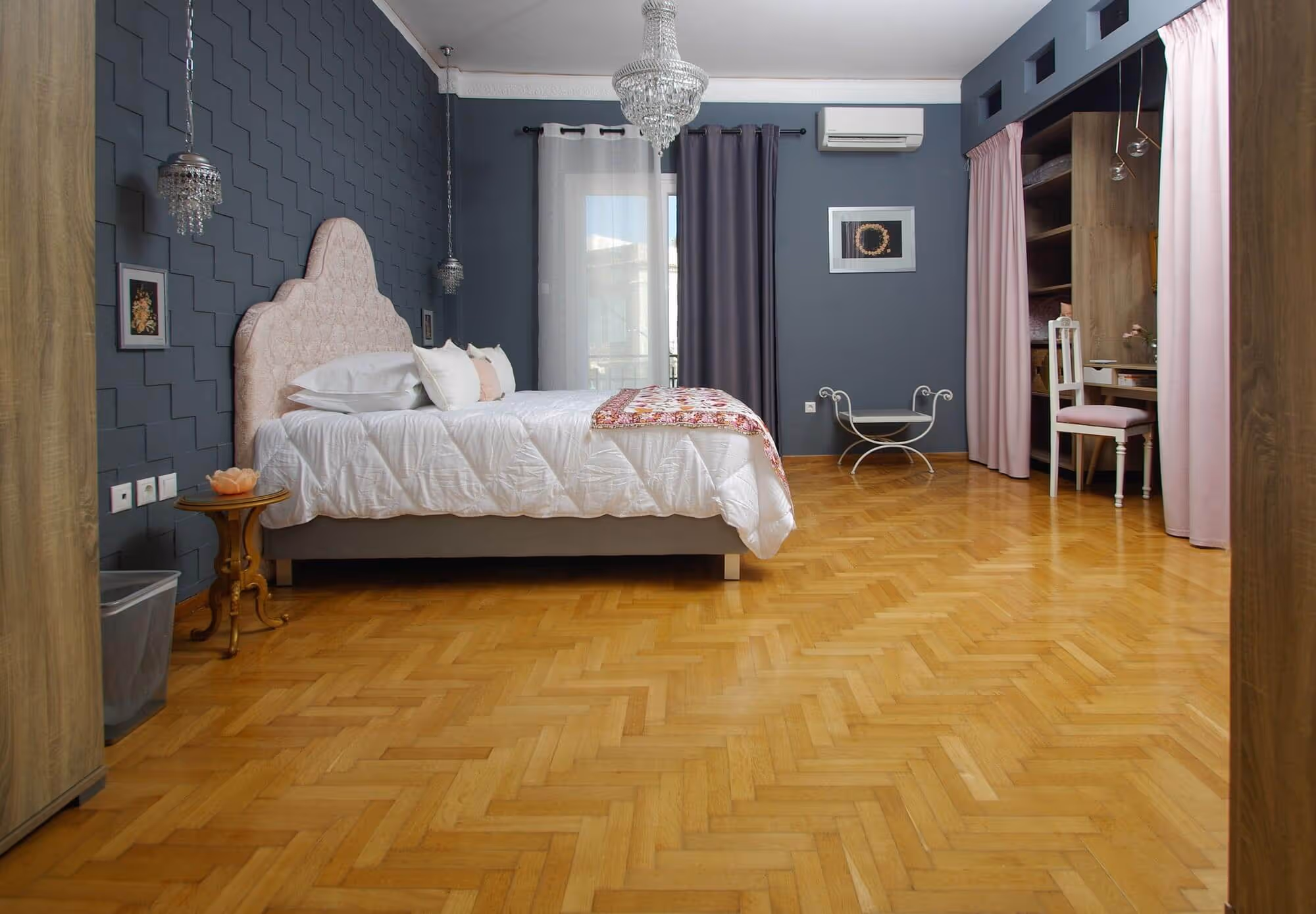 Spacious bedroom with wooden parquet flooring, a bed with a pink upholstered headboard, blue walls, and a chandelier.
