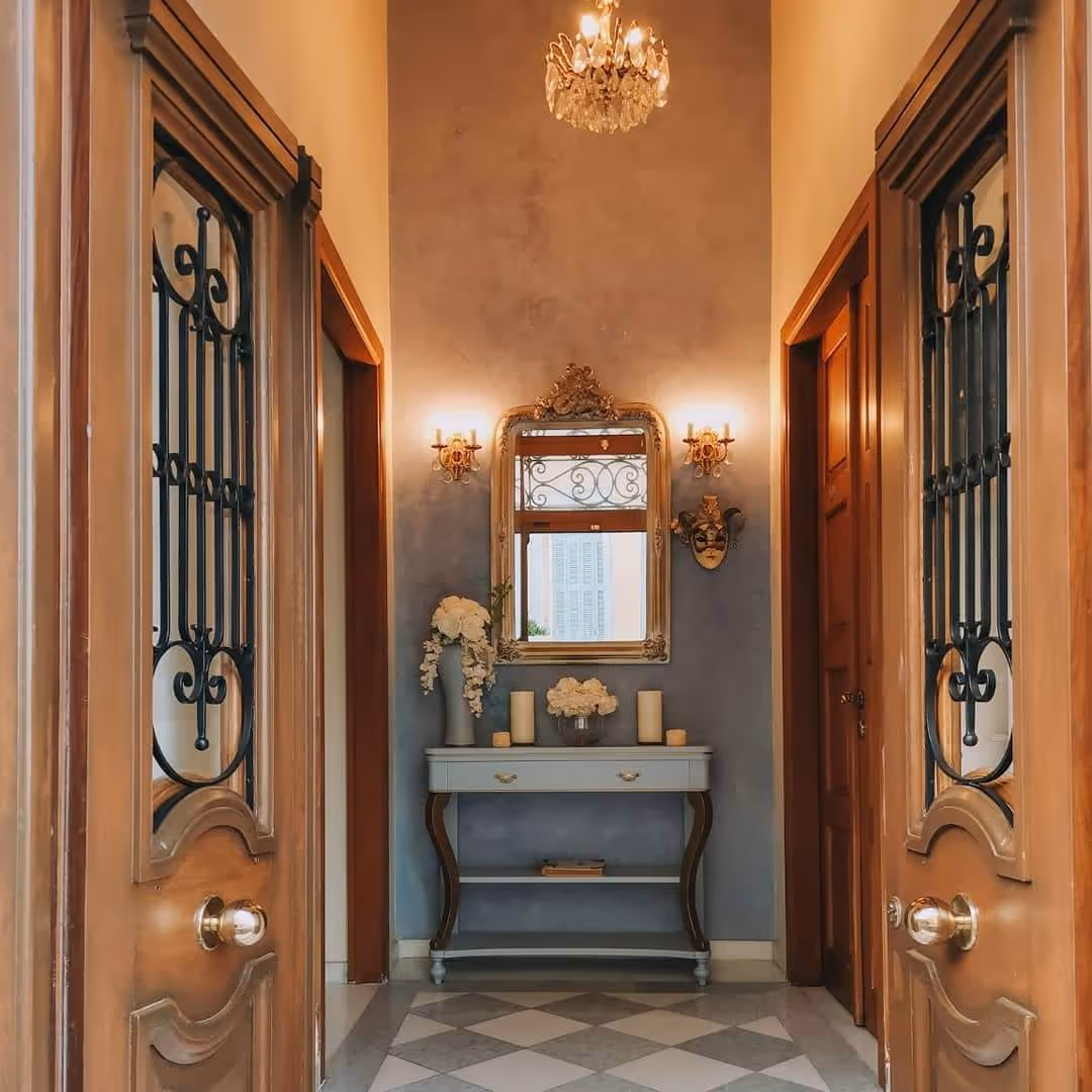 Elegant hallway with wooden doors, chandelier, ornate mirror, and a console table with flowers.