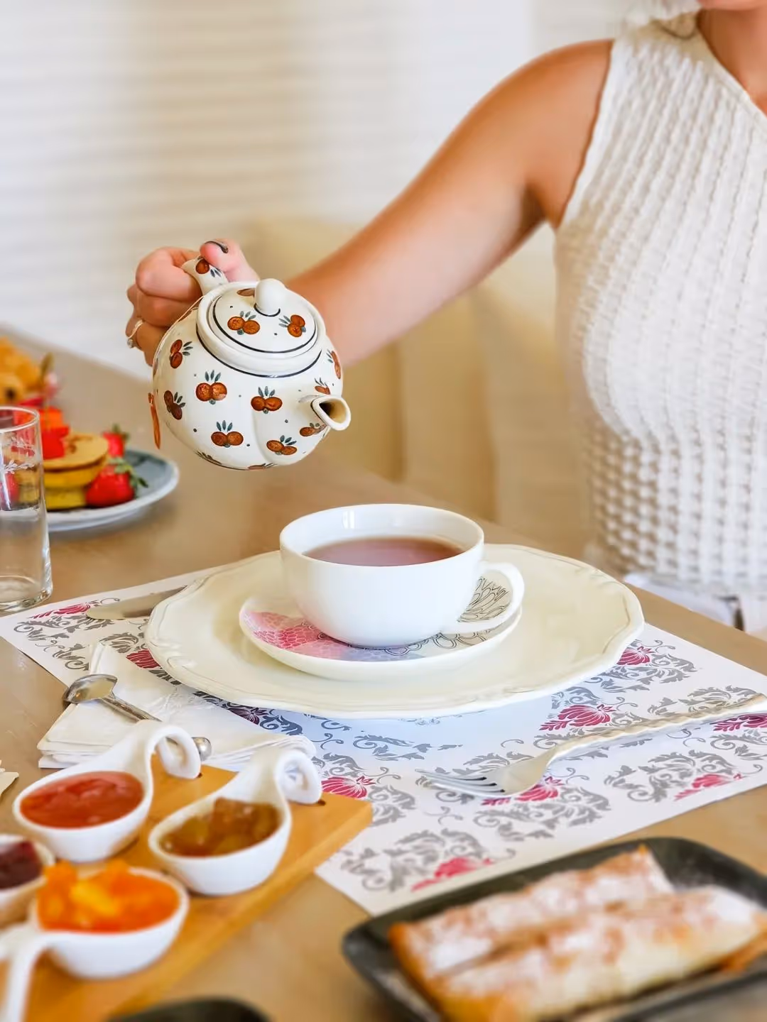 Person in white top pouring tea from a white teapot with orange fruit designs into a white cup.