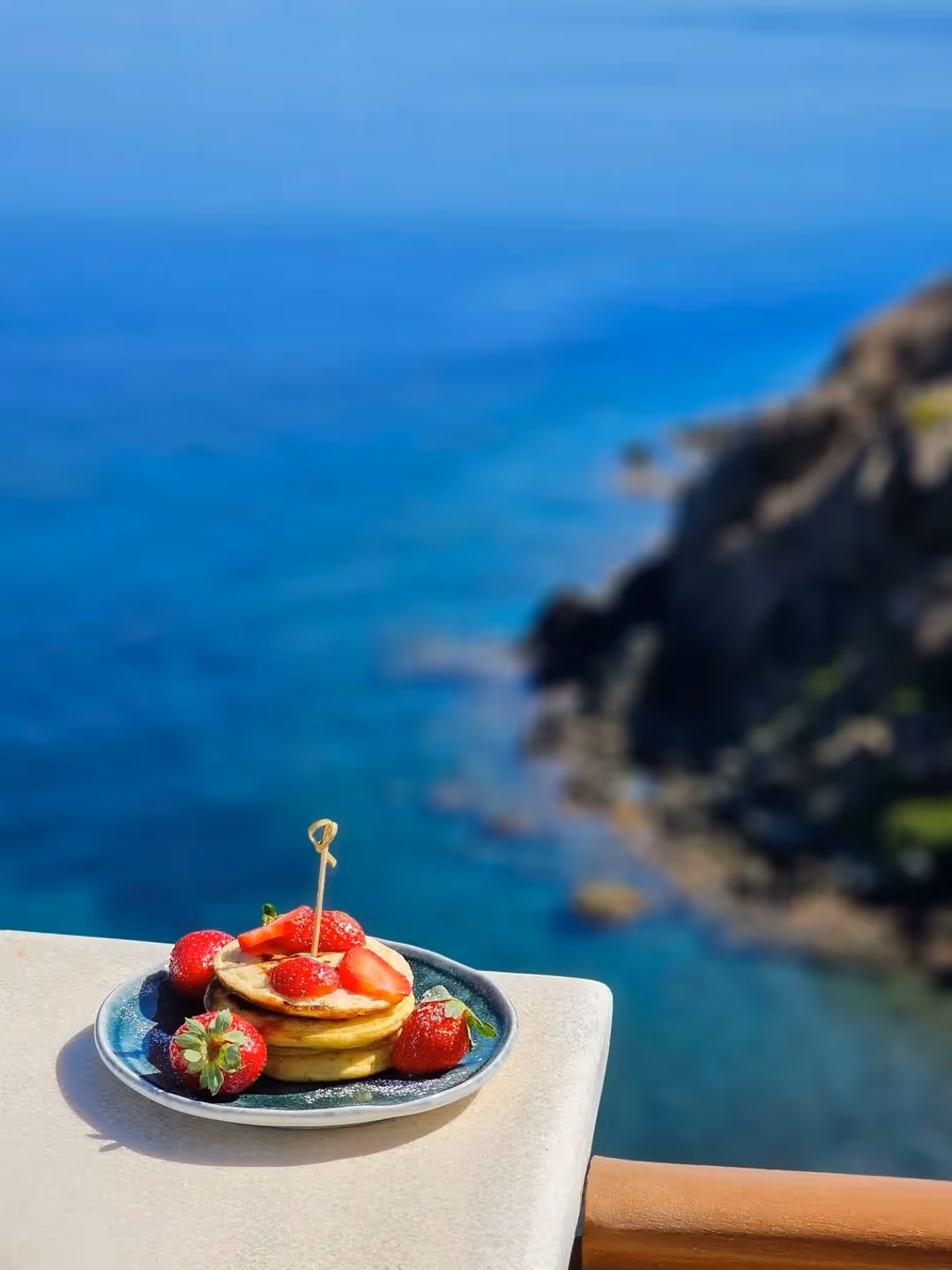 Stack of pancakes with fresh strawberries on a plate on a balcony overlooking a blue ocean and rocky coastline.