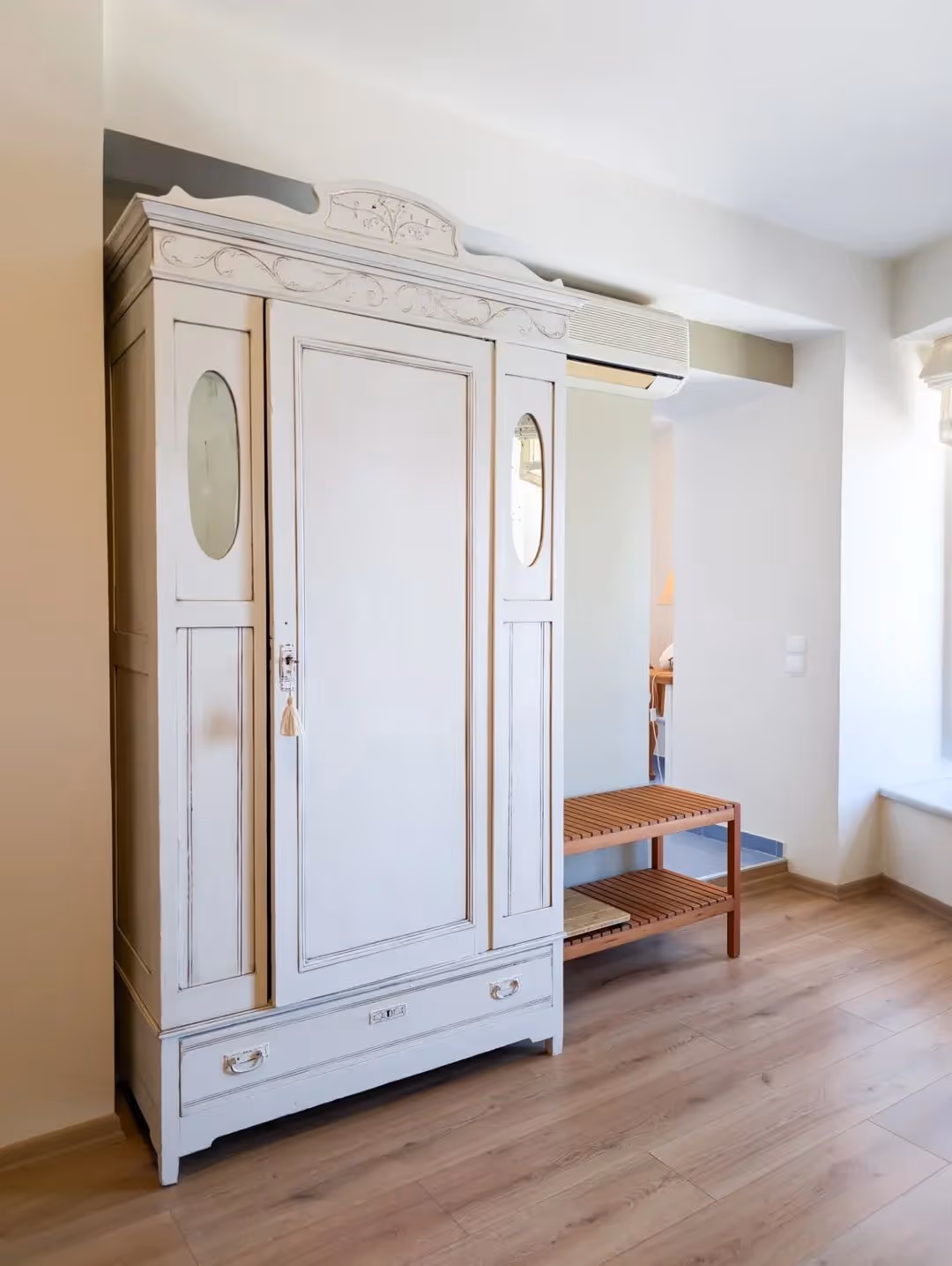 White vintage wardrobe with decorative carvings and oval mirrors next to a wooden slatted bench.