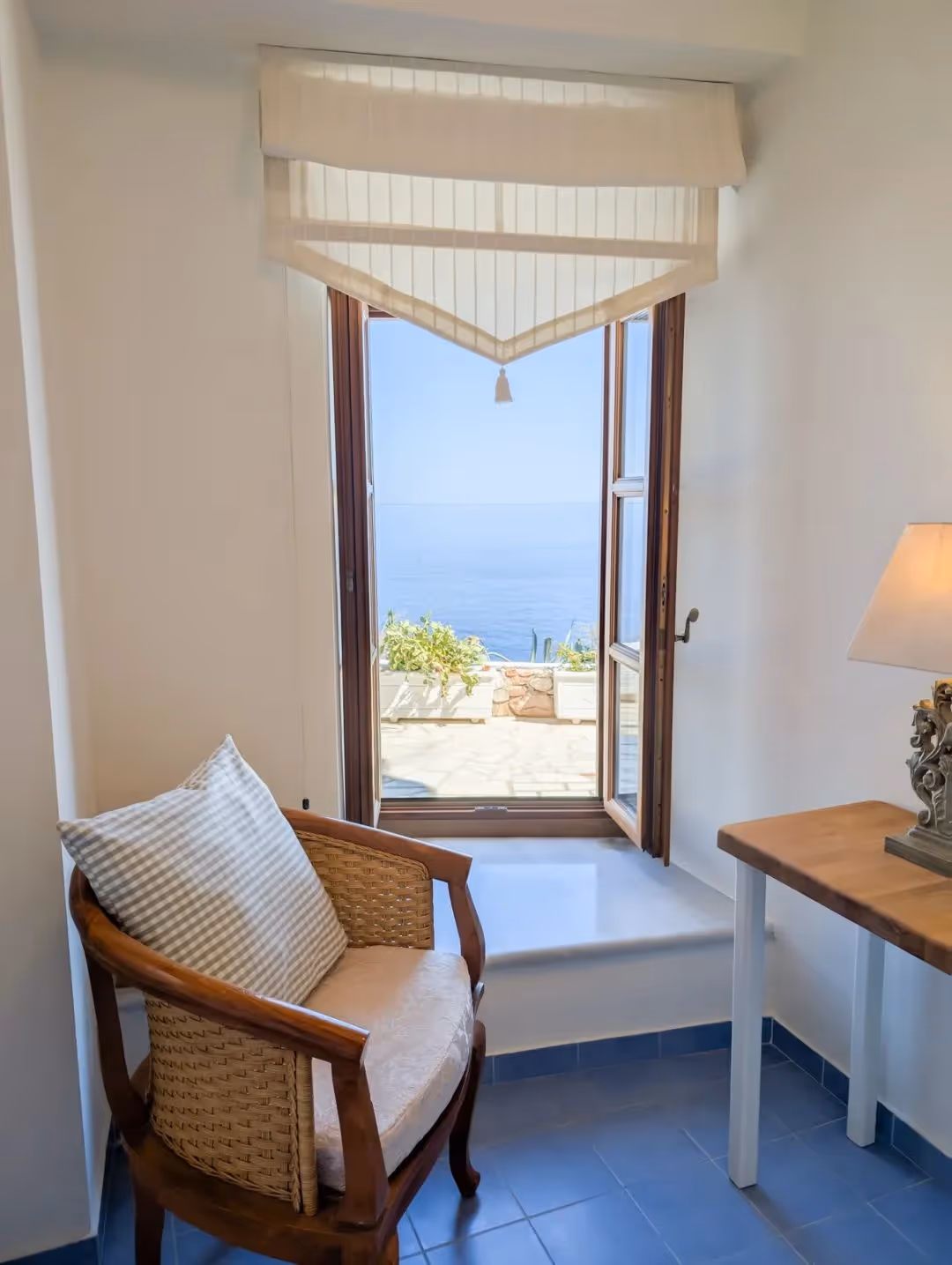 Cozy chair with cushion near open window showing sea view and sunlight outside.