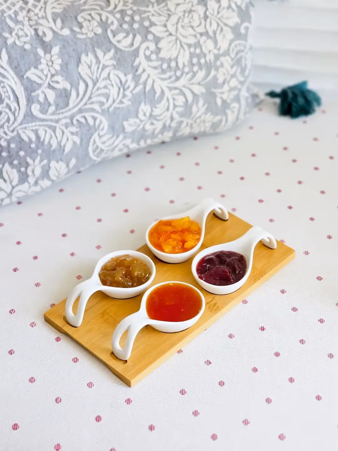 Four white ceramic spoons with different colored jams on a wooden board on a patterned cloth.