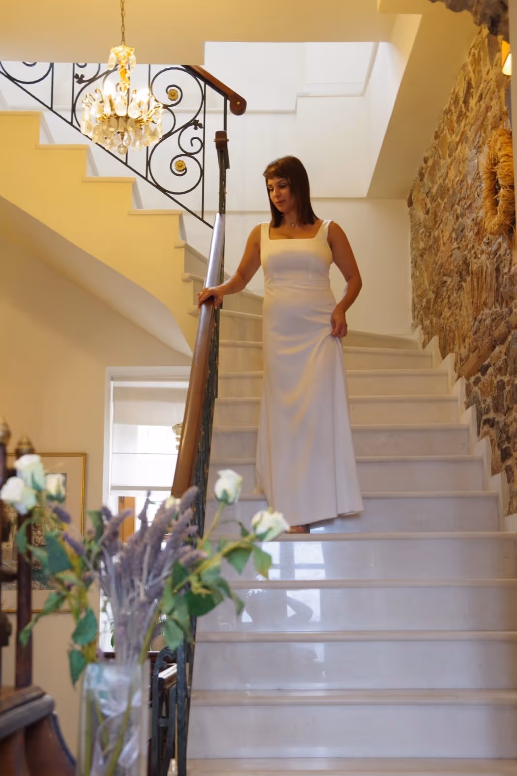 Woman in a white sleeveless dress descending an elegant marble staircase with a wooden and wrought iron railing.