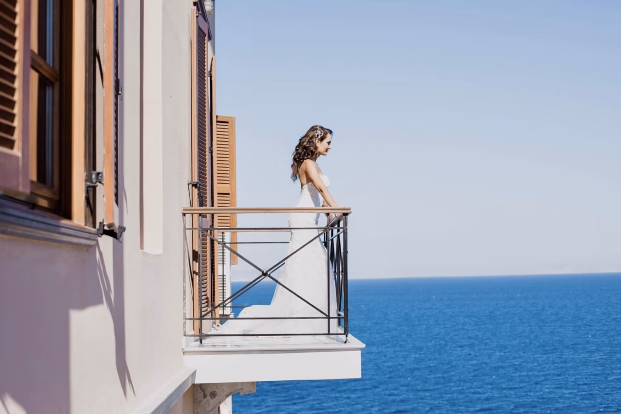 Bride in a white wedding dress standing on a balcony overlooking the blue ocean under a clear sky.