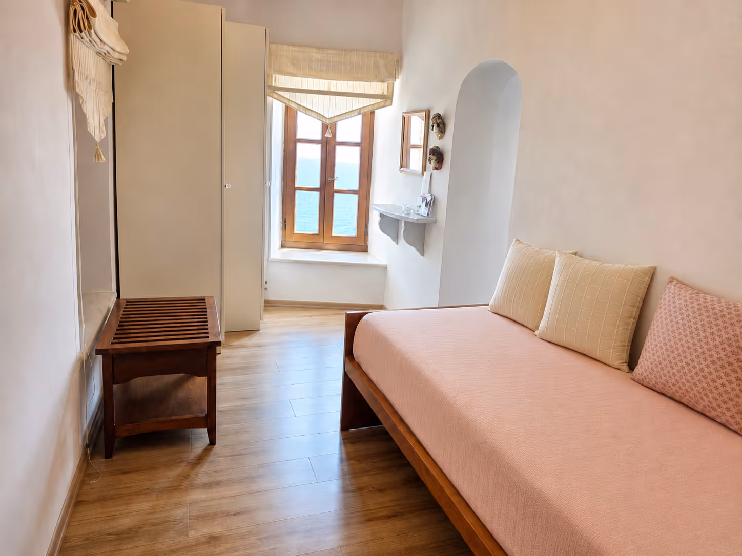 Minimalist room with a wooden daybed with cushions, a window showing sea view, and wooden floor.