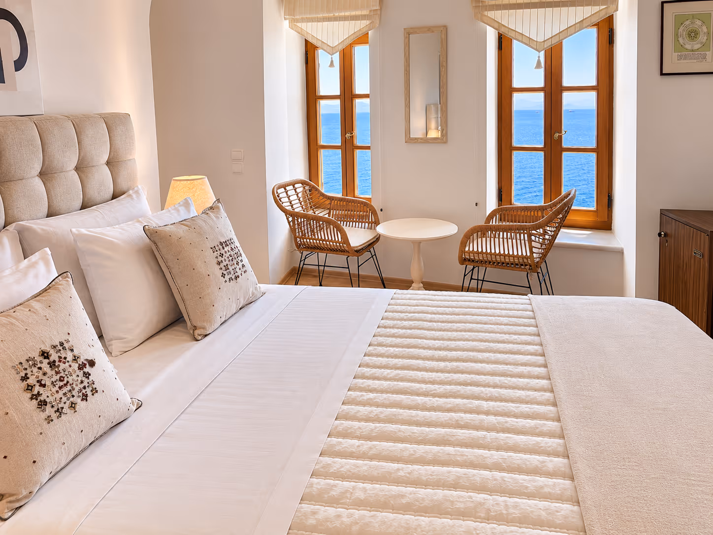 Cozy bedroom with beige bedding, pillows, two wicker chairs, small table, and ocean view through windows.