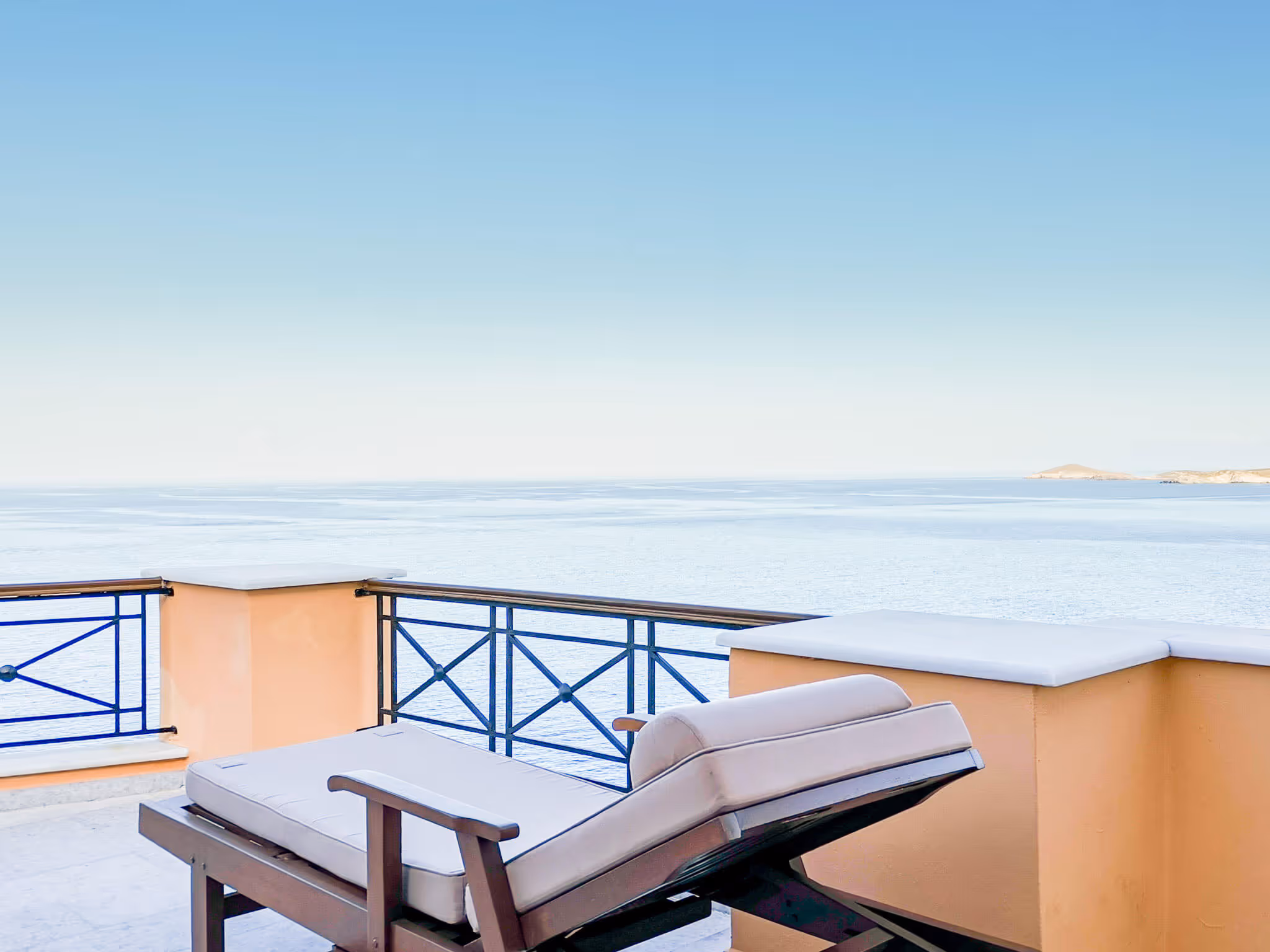 Cushioned lounge chair on a balcony overlooking calm blue sea and clear sky.