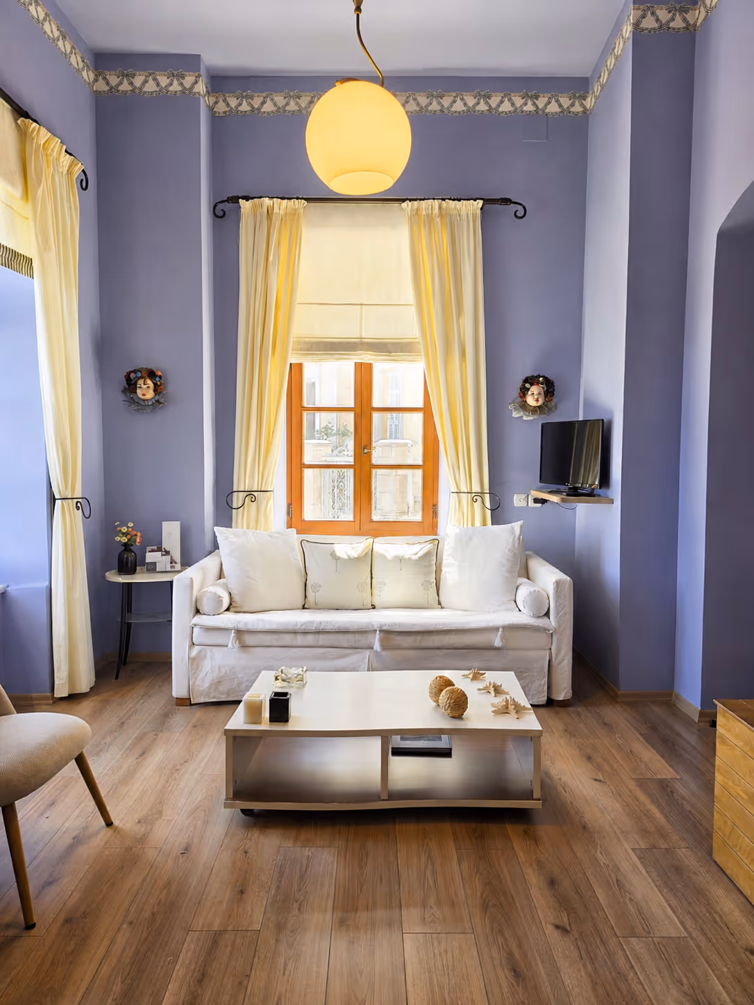 Living room with lavender walls, wooden floor, white sofa, coffee table, yellow curtains, and wall-mounted TV.