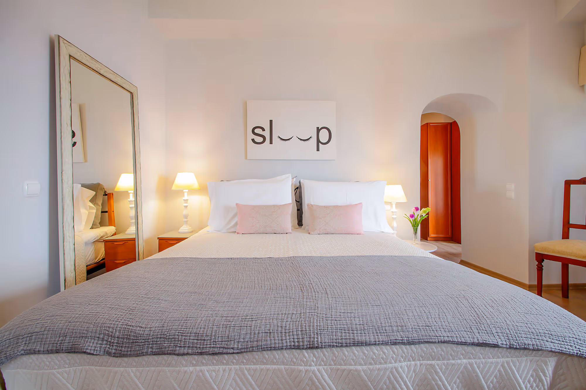 Bright bedroom with neatly made bed, two bedside lamps, decorative pillows, and a wall art reading 'sleep' with eyelash graphics.