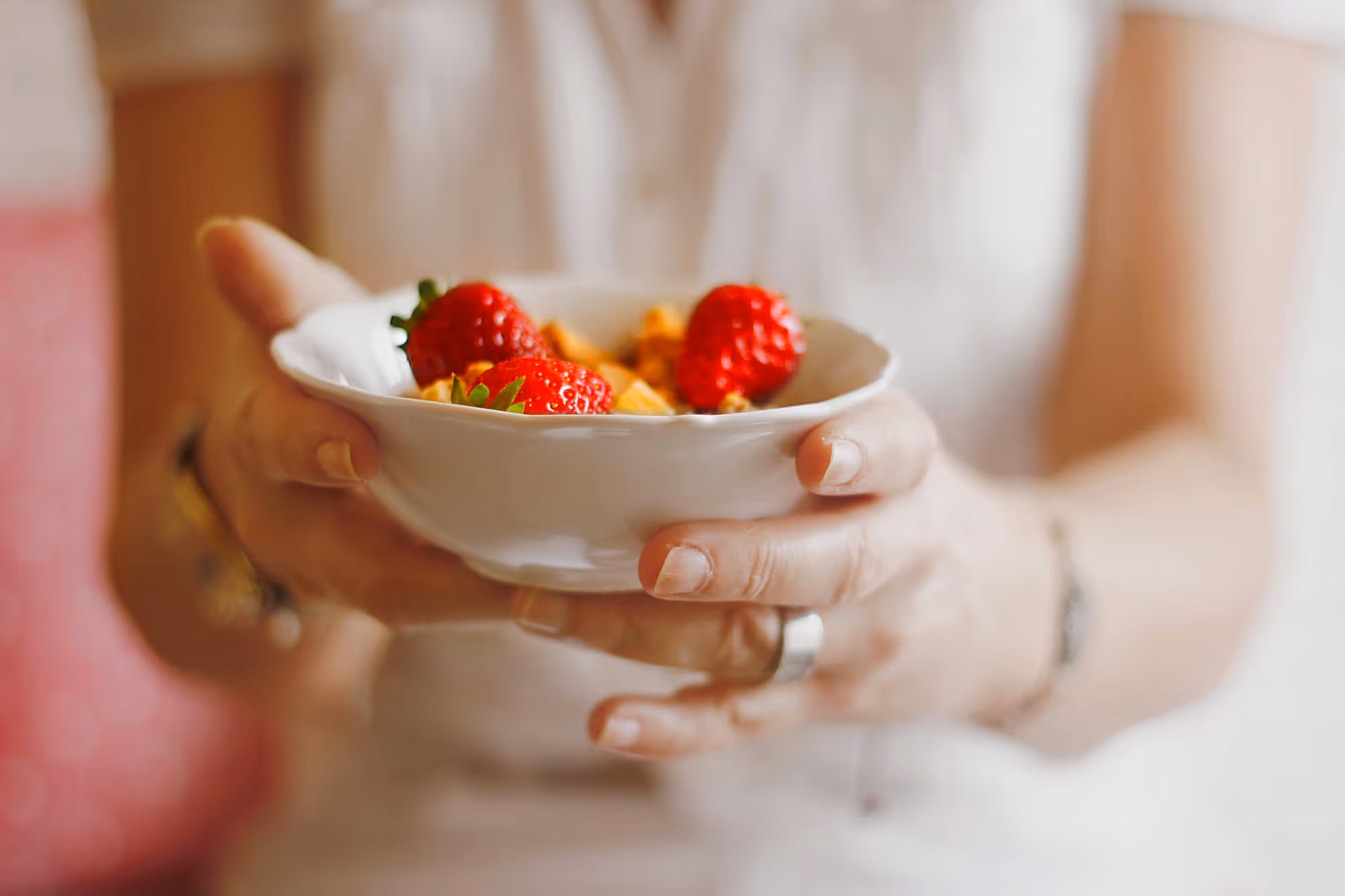 Hands holding a white bowl with strawberries and granola.