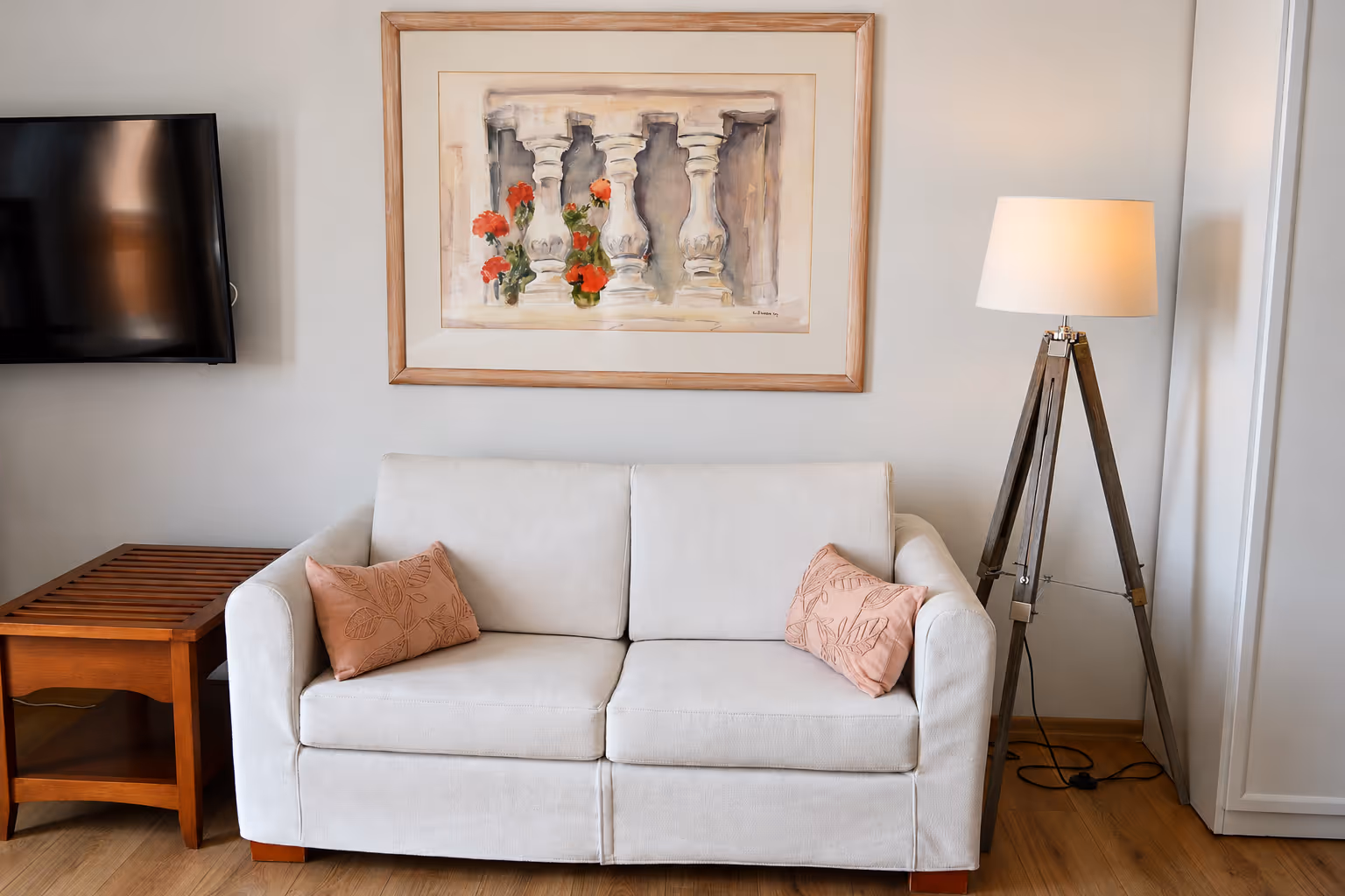 White sofa with two pink leaf-patterned pillows, wooden side table, tripod floor lamp, and framed art above.