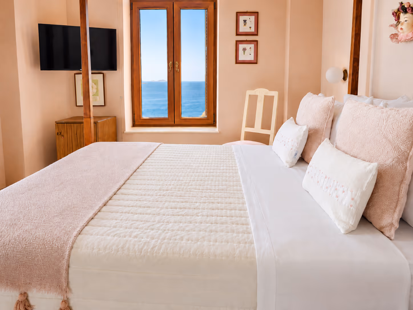 Cozy bedroom with a large bed, pink pillows, a window with ocean view, and a wall-mounted TV.