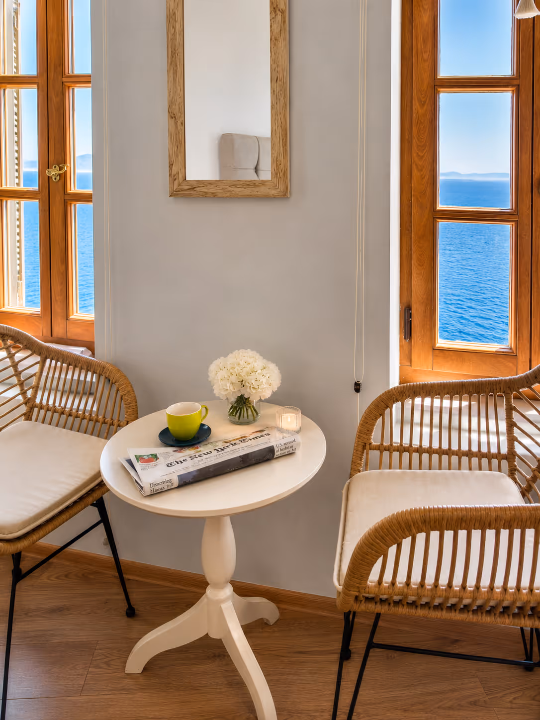 Cozy corner with rattan chairs, round white table, green cup, newspaper, white flowers, and ocean view.
