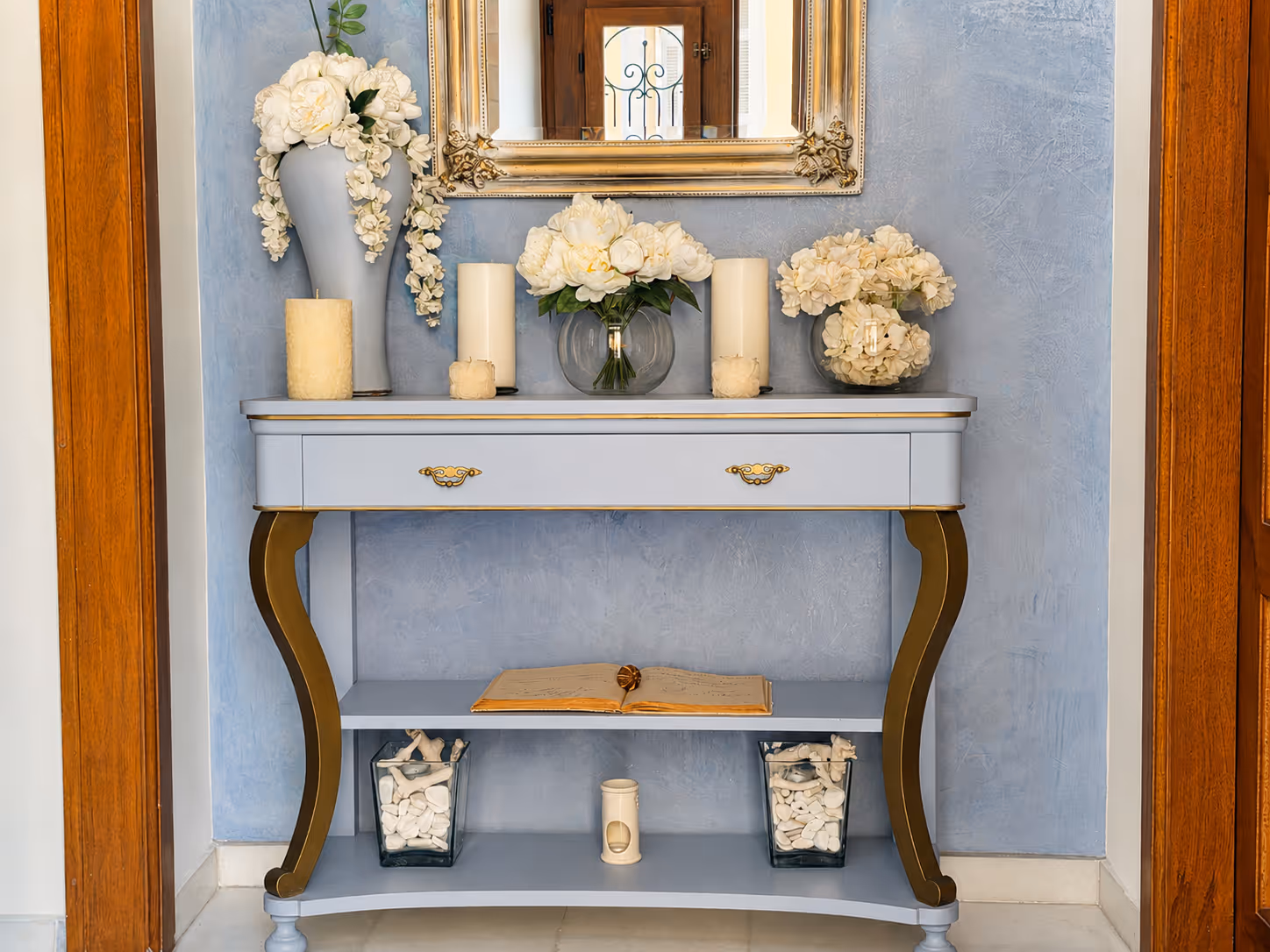 Blue console table with gold accents holding white flowers, candles, open book, and decorative stones.