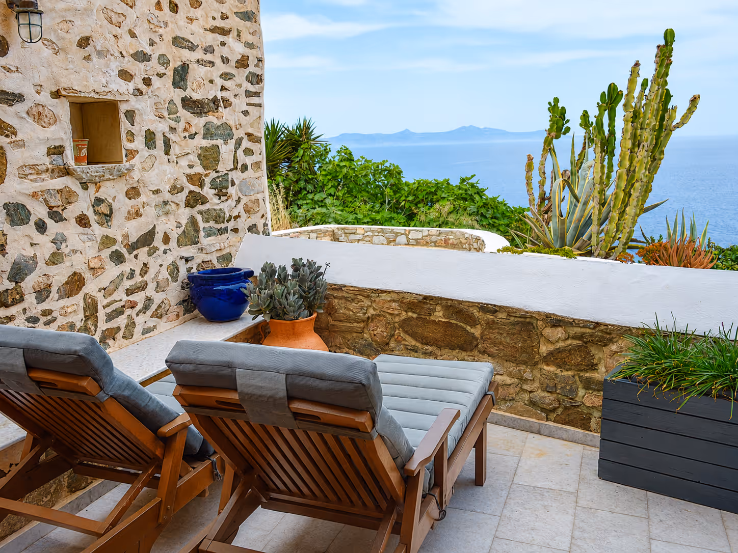 Two cushioned wooden lounge chairs on a stone terrace with cacti and ocean view.