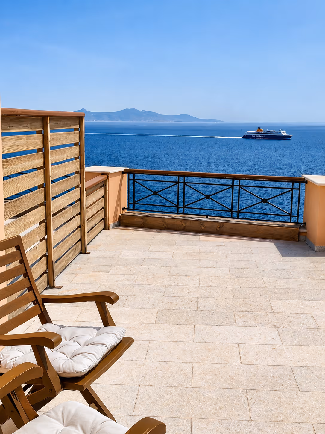 Balcony with wooden chairs overlooking blue sea and a ferry with distant mountains under clear sky.