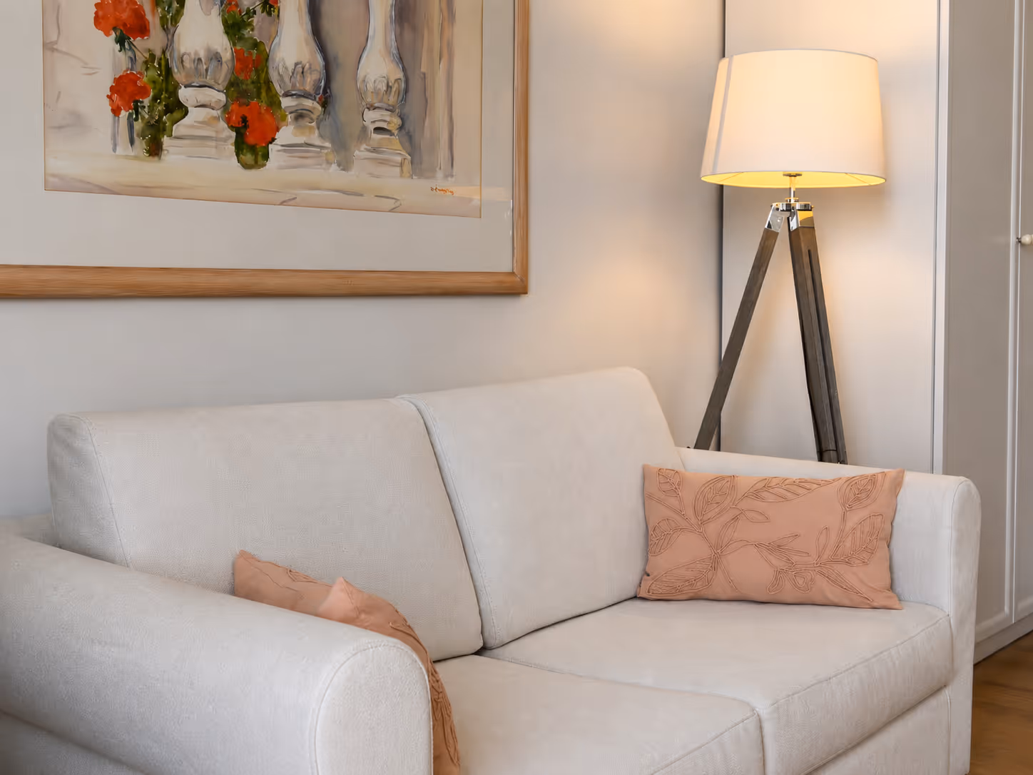 White sofa with two pink decorative pillows, standing next to a tripod floor lamp and framed wall art.