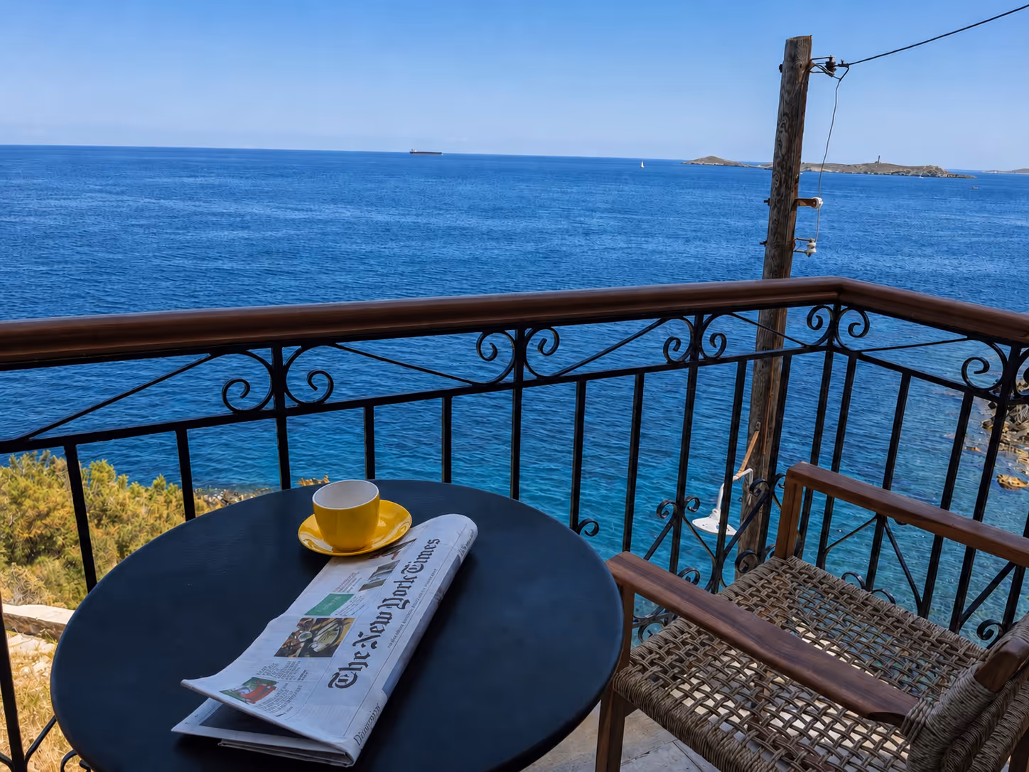 Balcony with a round table holding a yellow cup and folded newspaper overlooking a blue sea view.