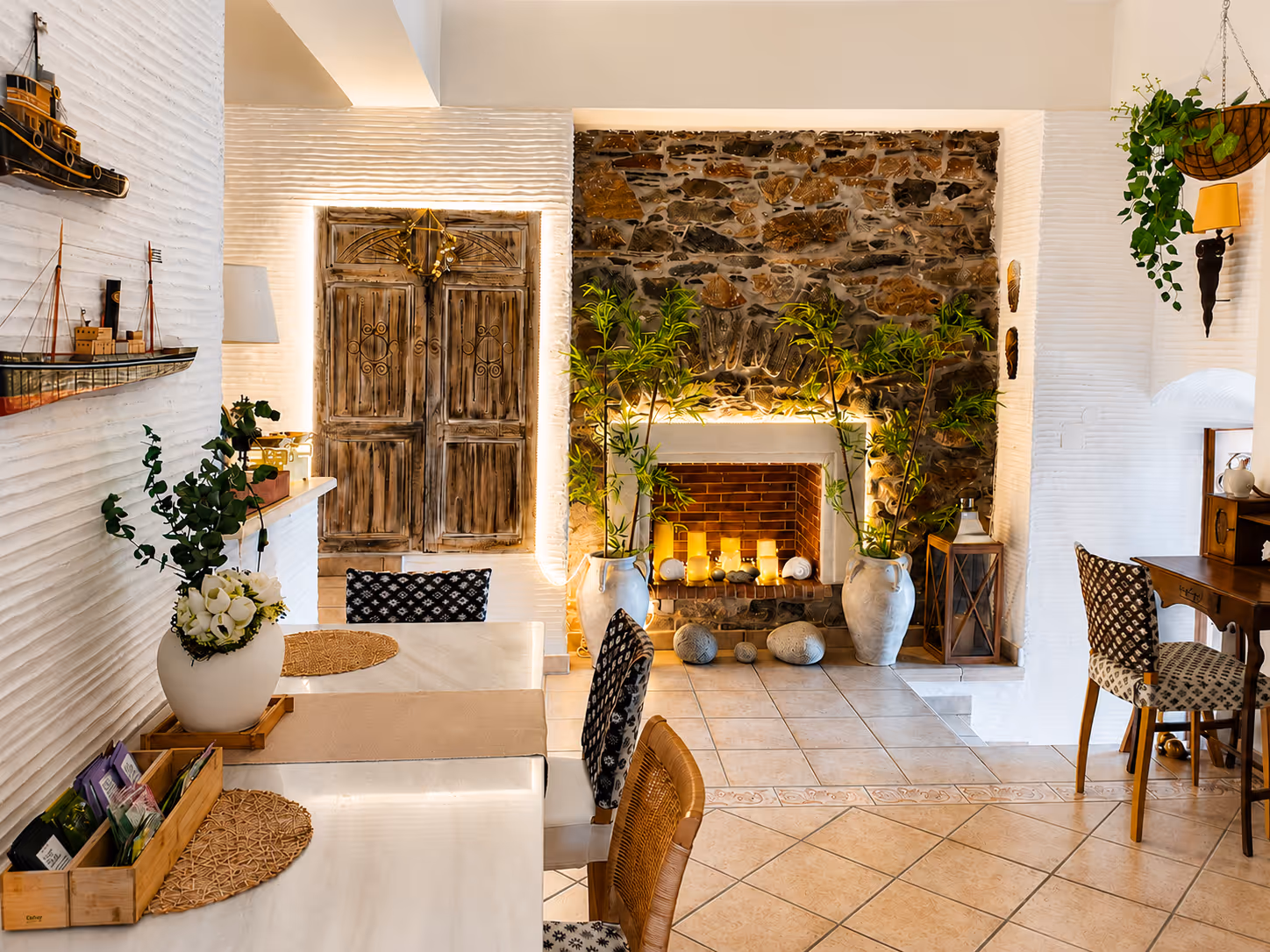 Cozy dining room with lit stone fireplace, rustic wooden doors, patterned chairs, and plants.
