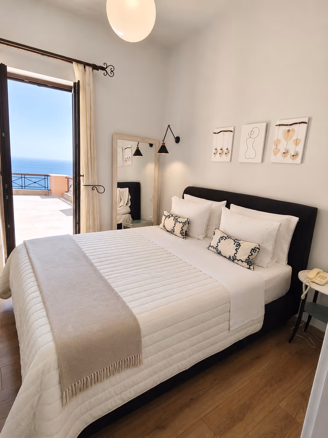 Bright bedroom with white bed, beige throw, pillows, wall decor, large mirror, and balcony with sea view.