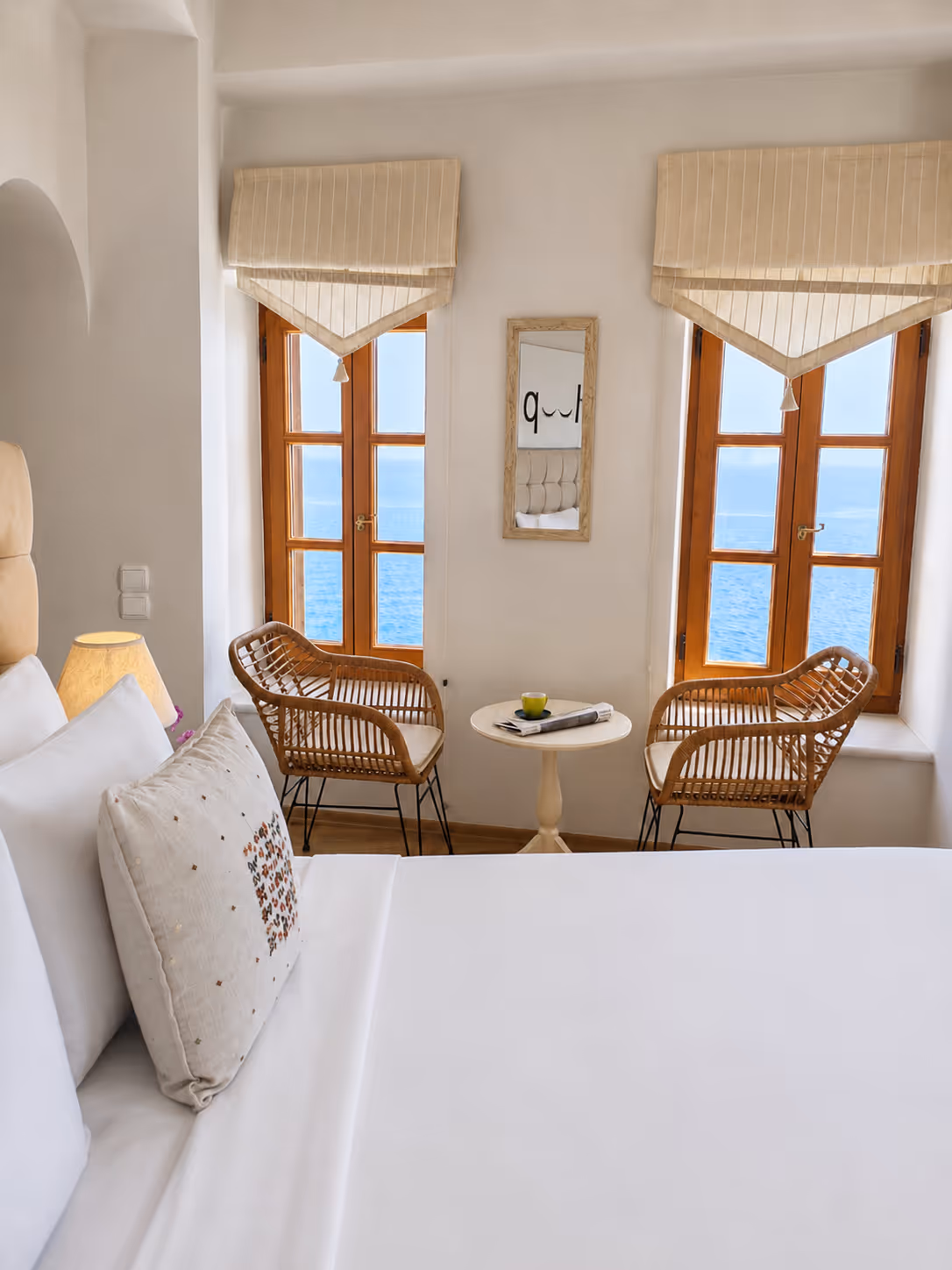 Bright bedroom with white bedding, two wicker chairs, a round table, and windows overlooking the sea.