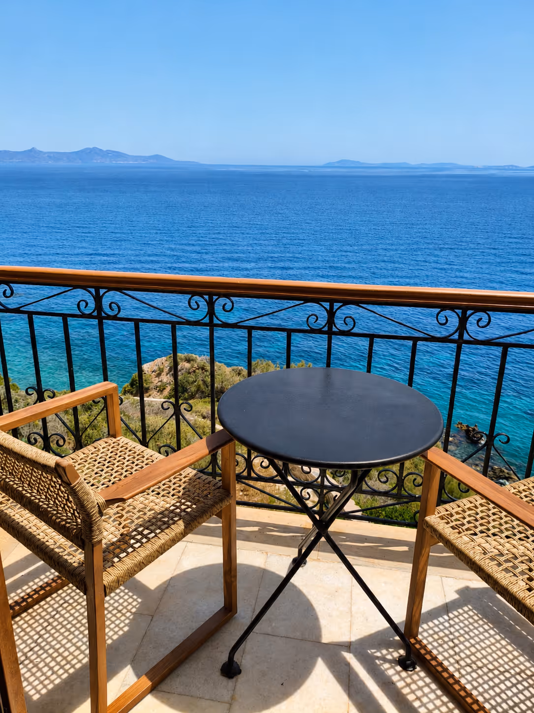 Two woven chairs and a small round table on a balcony overlooking blue ocean and distant mountains.