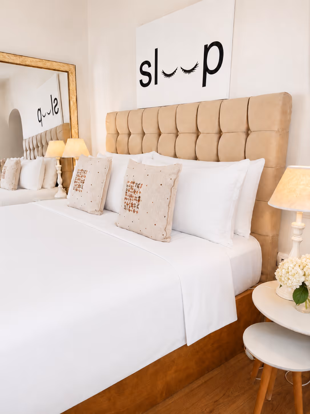 Bed with beige tufted headboard, white bedding, decorative pillows, lamps, and a mirror reflecting the room.
