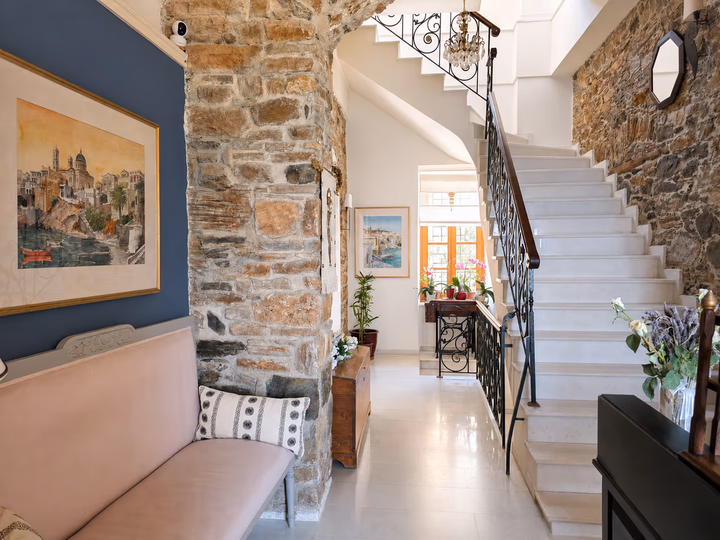 Bright hallway with stone walls, white staircase, cushioned bench, plants, and framed artwork.