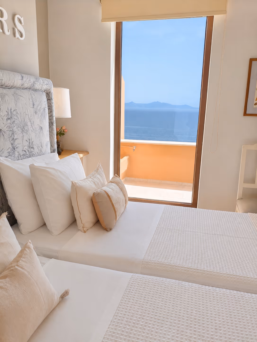 Two neatly made beds with white linens face a sliding door overlooking an ocean view.