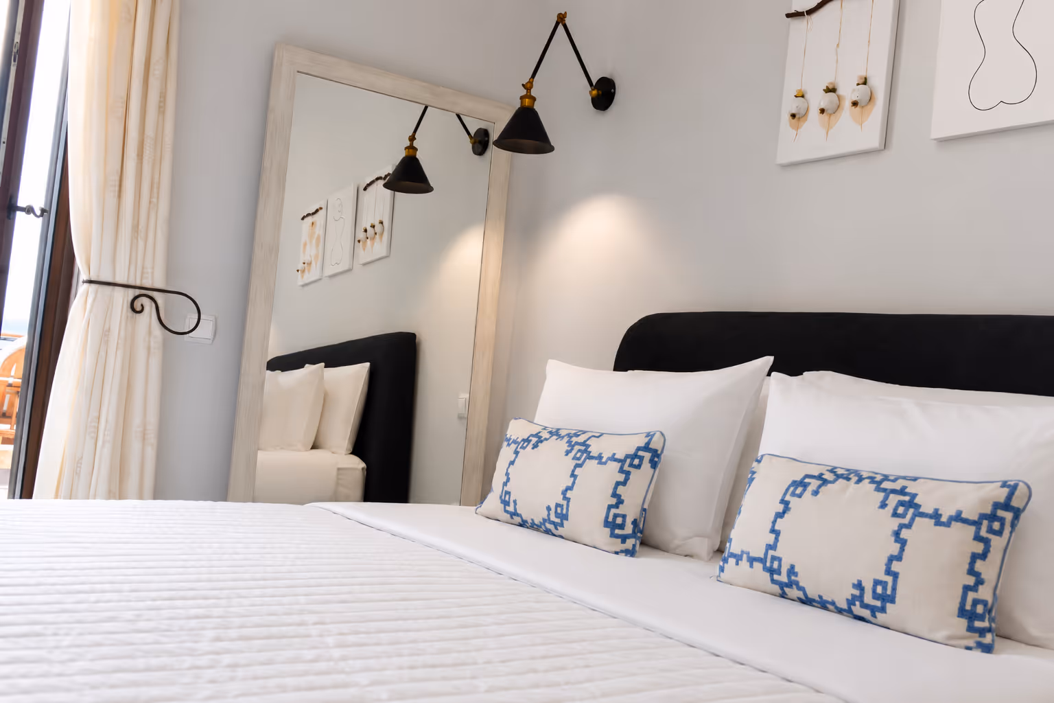 Bed with white linens, blue patterned pillows, black headboard, wall art, and a large mirror reflecting the bed.