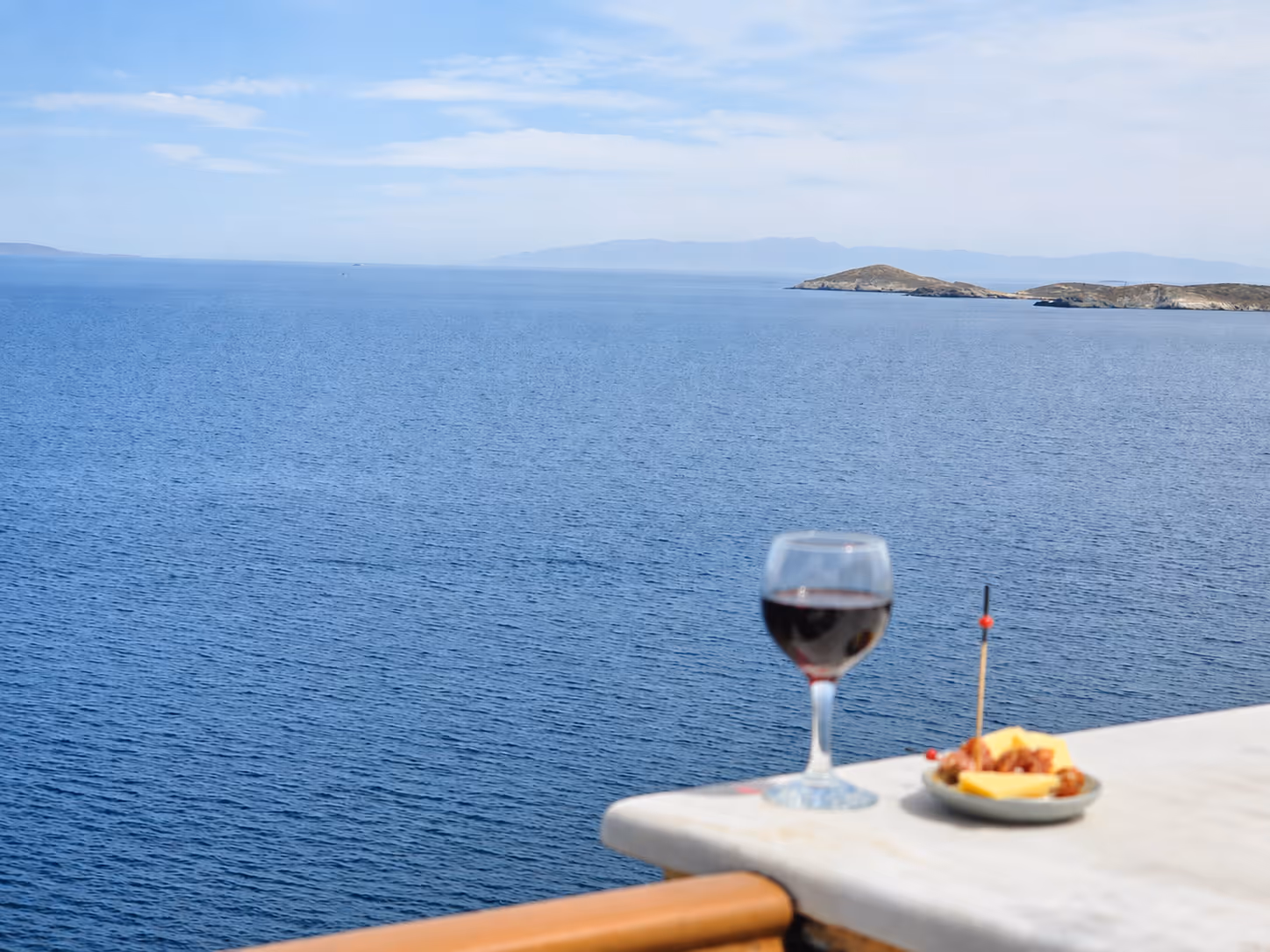 Glass of red wine and small plate with snacks on a table overlooking calm blue sea and distant islands.