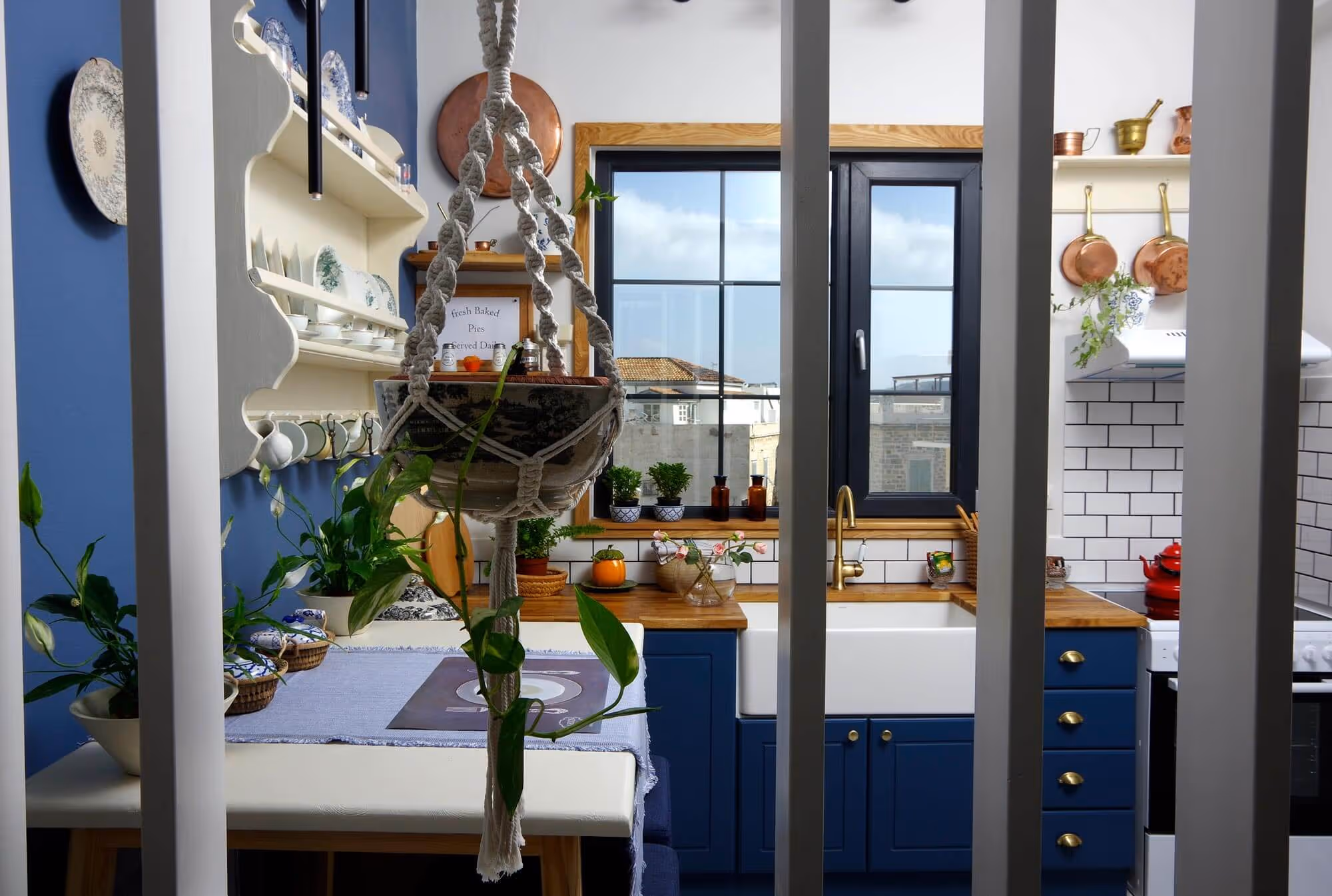 Cozy kitchen with blue cabinets, wooden countertops, white farmhouse sink, hanging plants, copper pots, and a window showing an outdoor view.