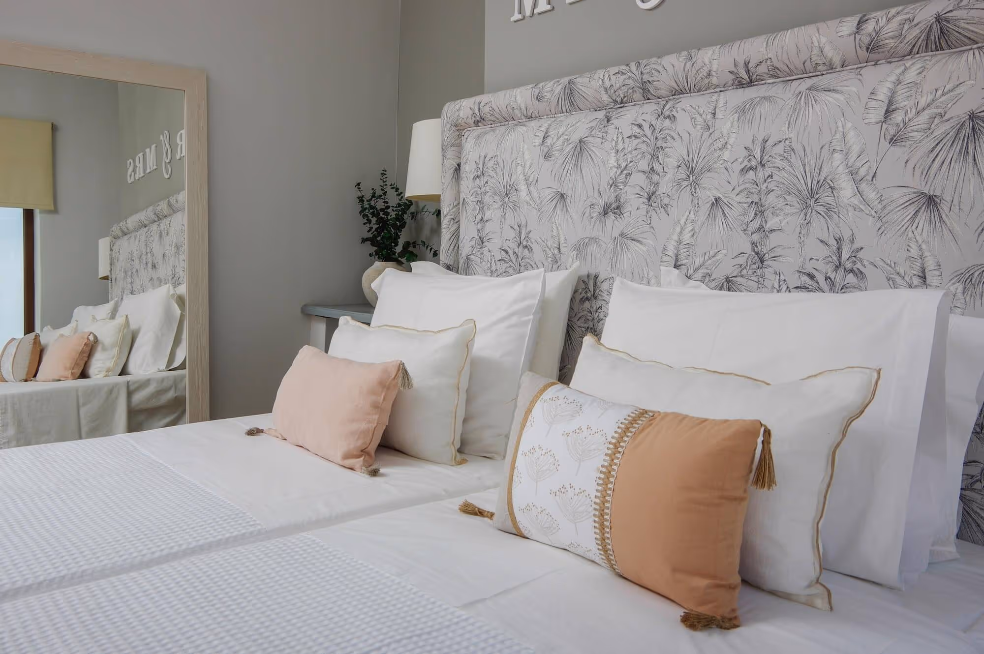 Bed with floral-patterned headboard, white bedding, and decorative pillows in cream and peach tones, reflected in a large mirror.