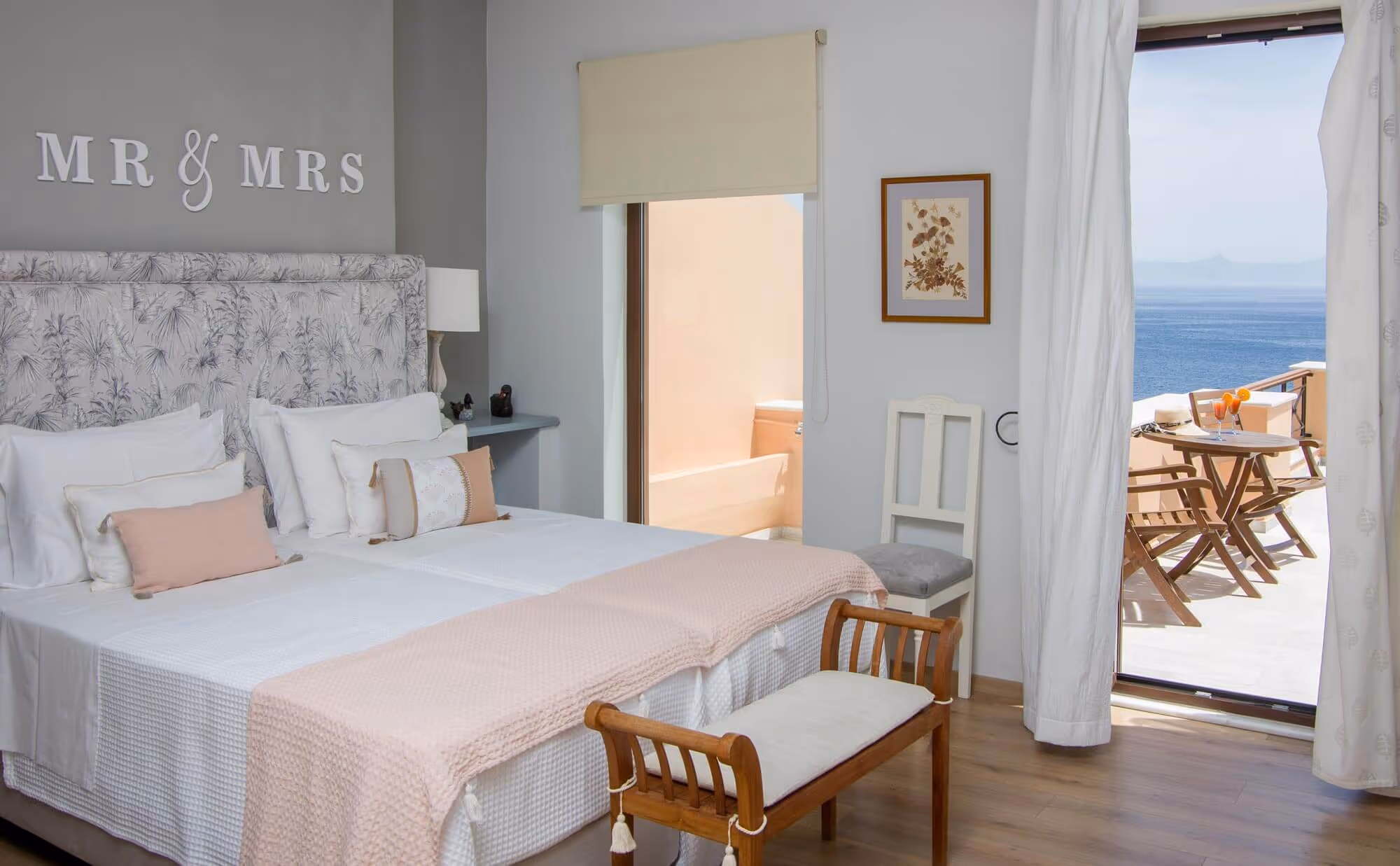 Bright bedroom with twin beds, soft pink and white bedding, a wooden bench, and a balcony overlooking a blue sea.