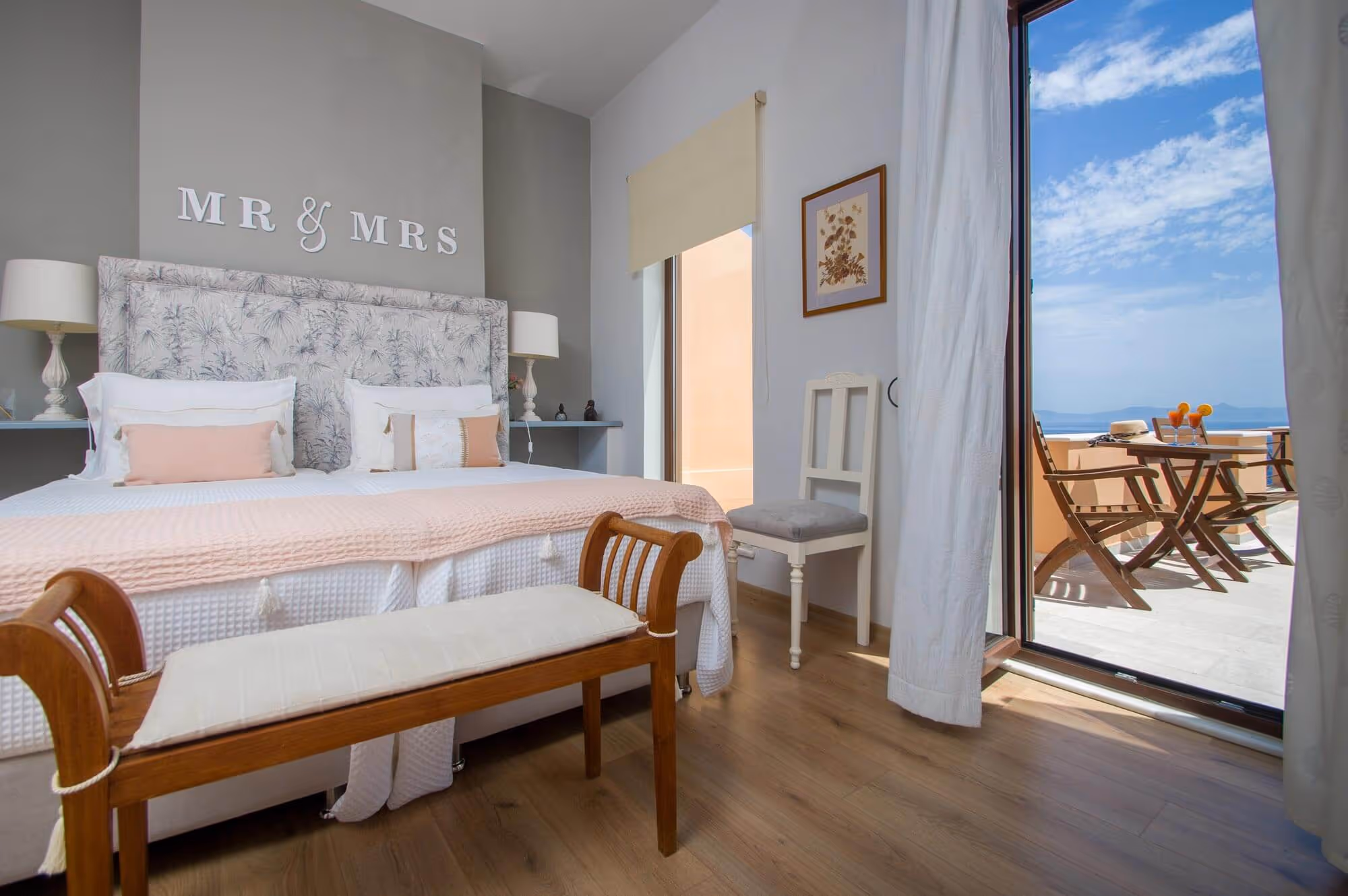 Bright bedroom with a double bed labeled MR & MRS, soft pink bedding, wooden bench, and an open door leading to a balcony with chairs and a table overlooking a blue sky.