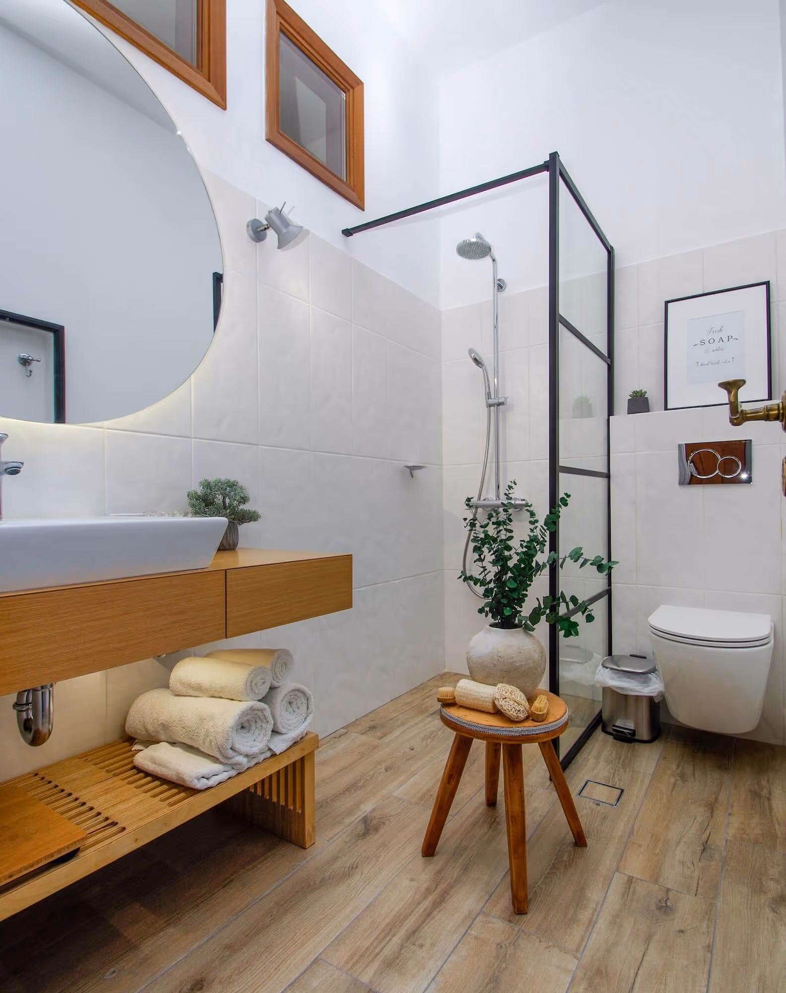 Modern bathroom with wooden floor, a floating wooden vanity holding a white sink and rolled towels, a glass-enclosed shower, a small wooden stool with bath brushes and a vase with green foliage, and a wall-mounted toilet.