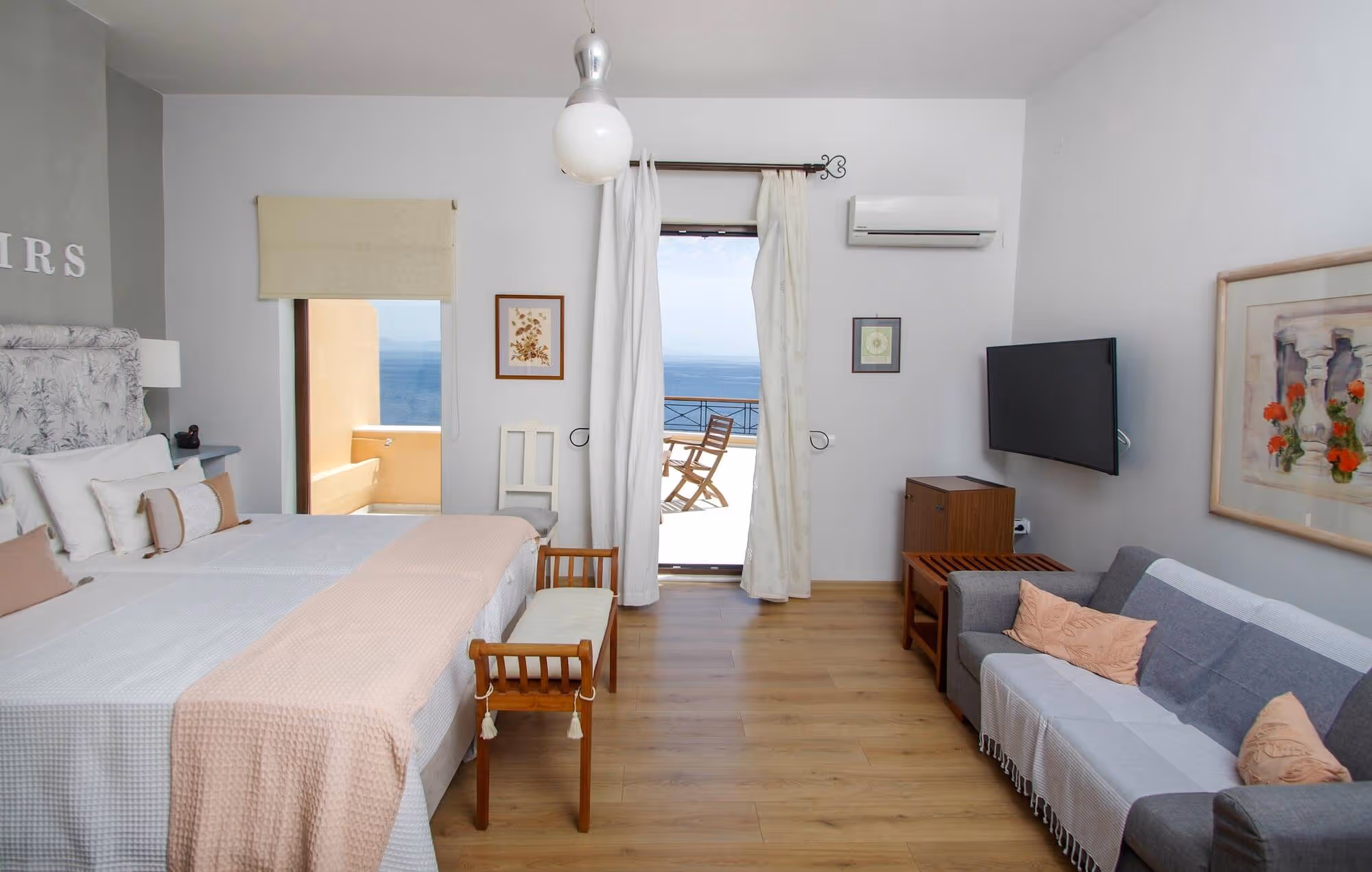 Bright hotel room with twin beds, a wooden bench, gray sofa with peach pillows, and a balcony with ocean view.