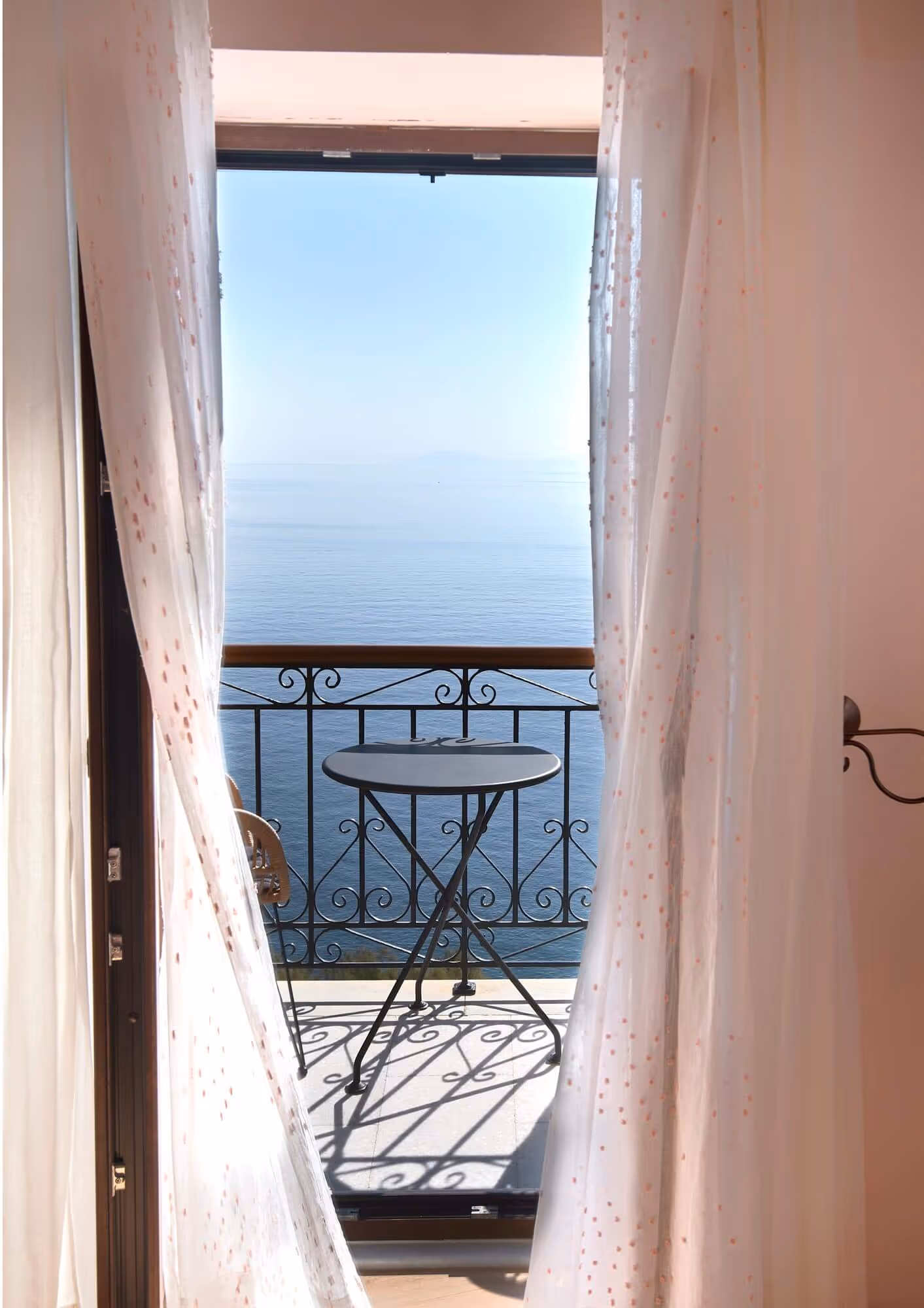 View through sheer curtains onto a balcony with a small table and chair overlooking a calm sea.