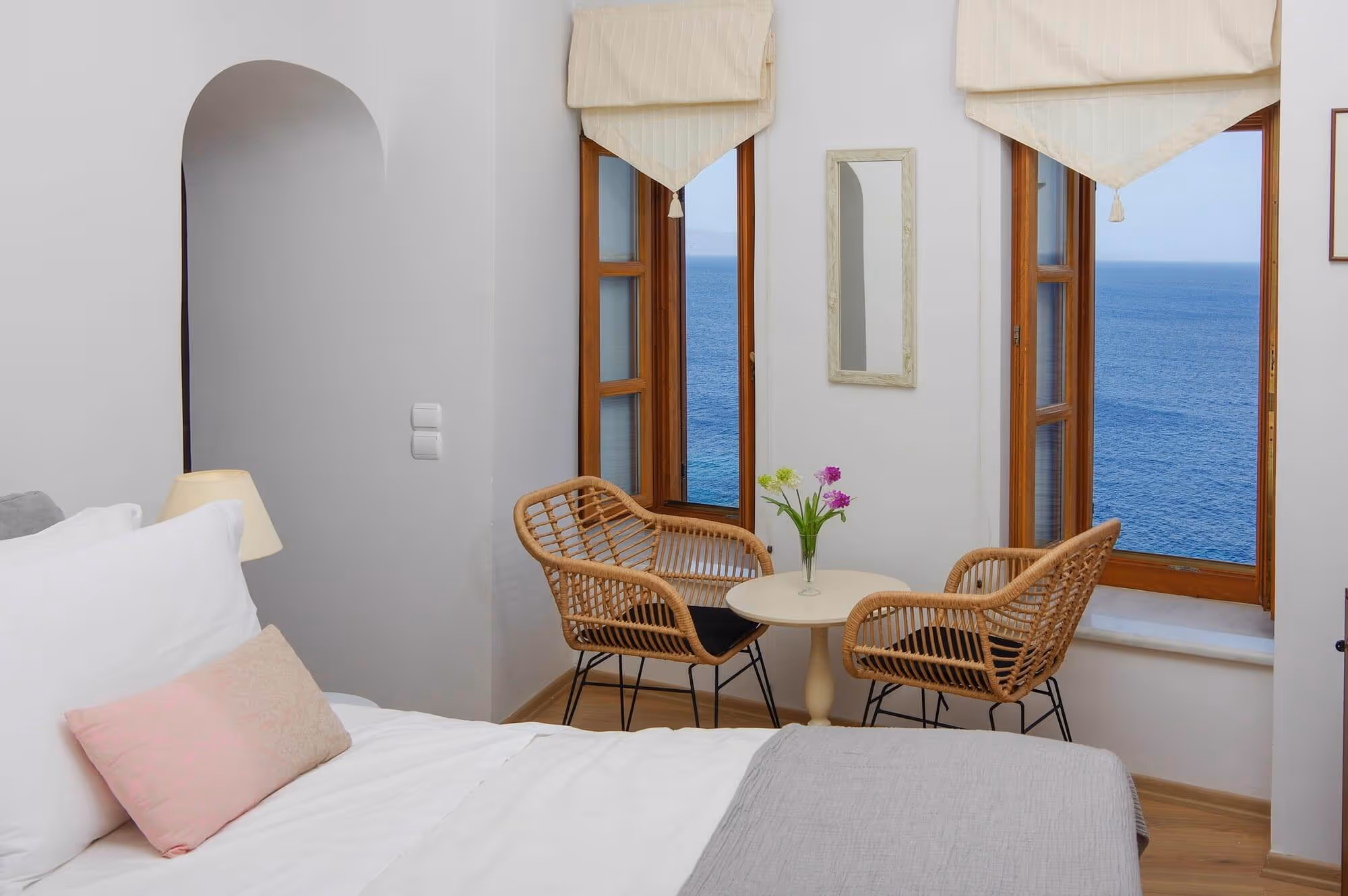 Cozy bedroom corner with white bed, two wicker chairs, small round table with flowers, and windows overlooking the ocean.
