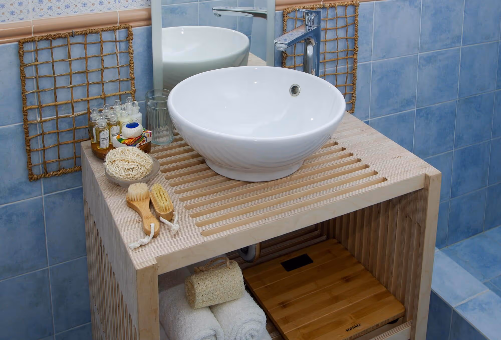 Bathroom countertop with a white vessel sink, chrome faucet, wooden brushes, soap, and toiletries on a light wooden slatted stand with towels and a weight scale underneath.
