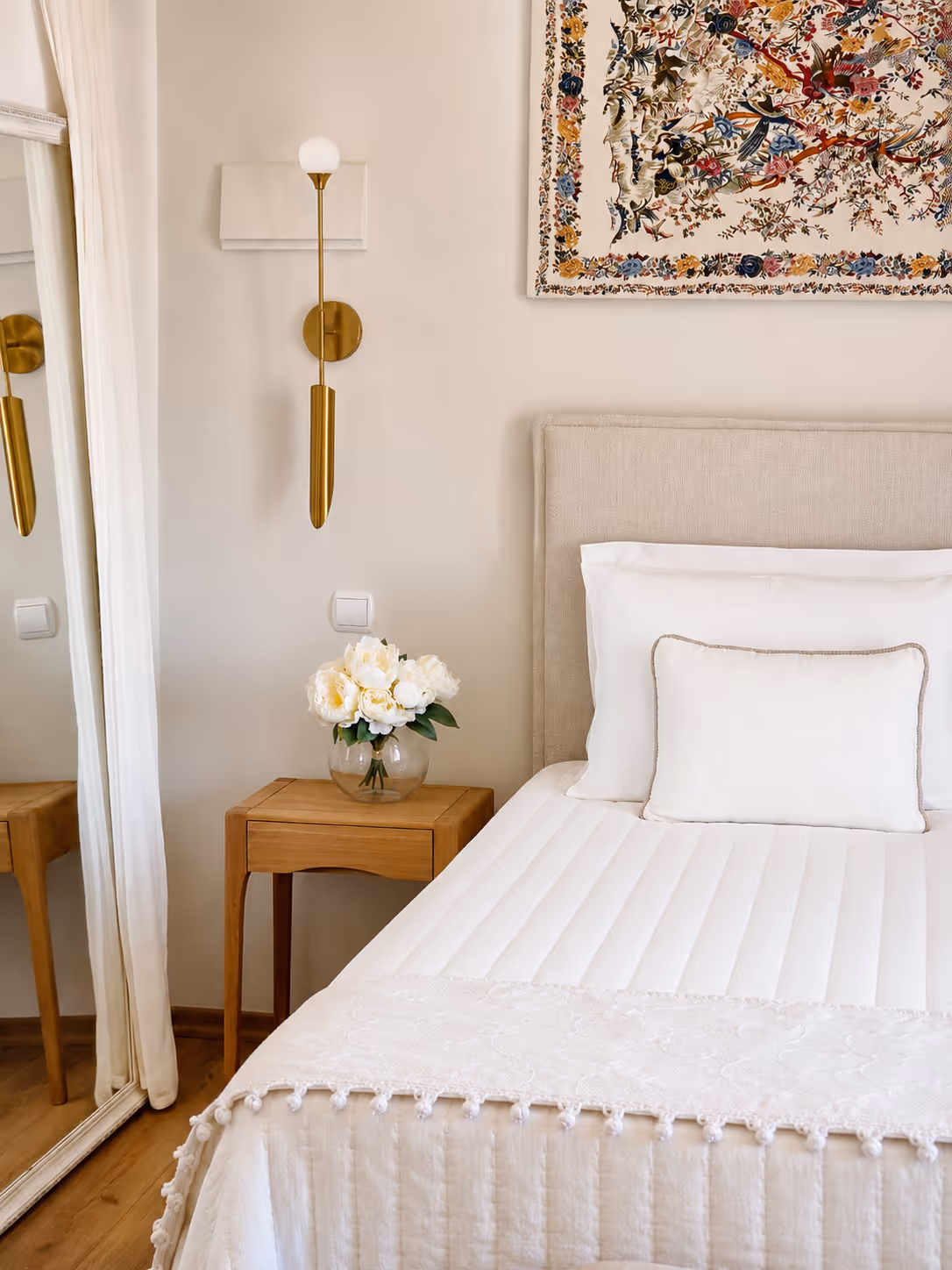 Neatly made bed with white linens and a small wooden nightstand holding a vase of white flowers.