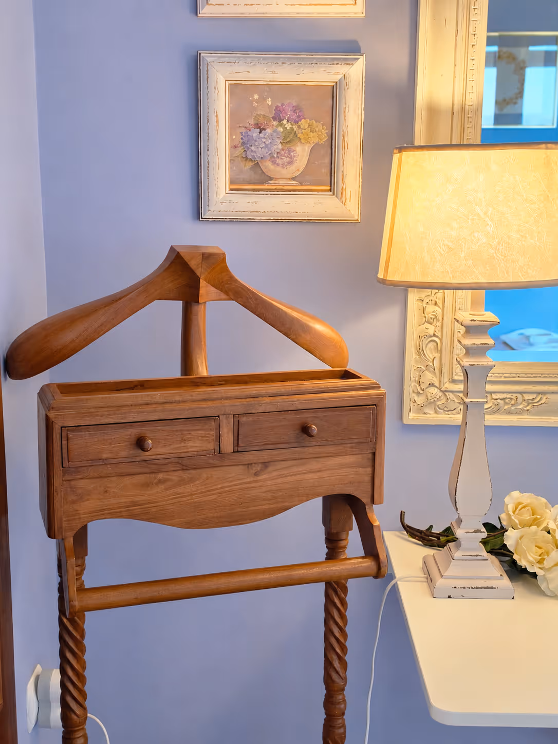 Wooden valet stand with drawers next to a white table holding a lit lamp and yellow roses.