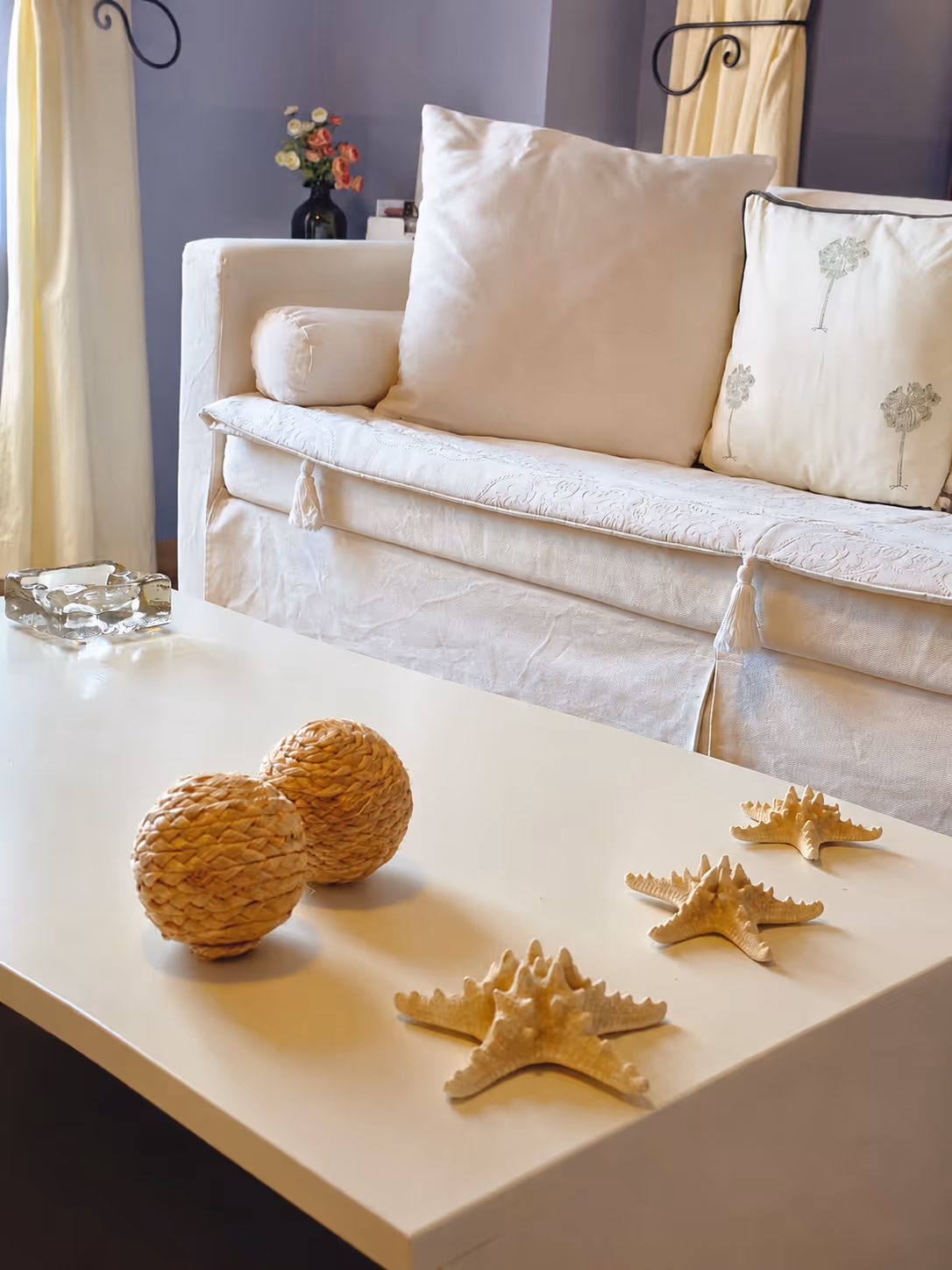 White coffee table with decorative woven balls and starfish figurines in front of a beige couch with cushions.