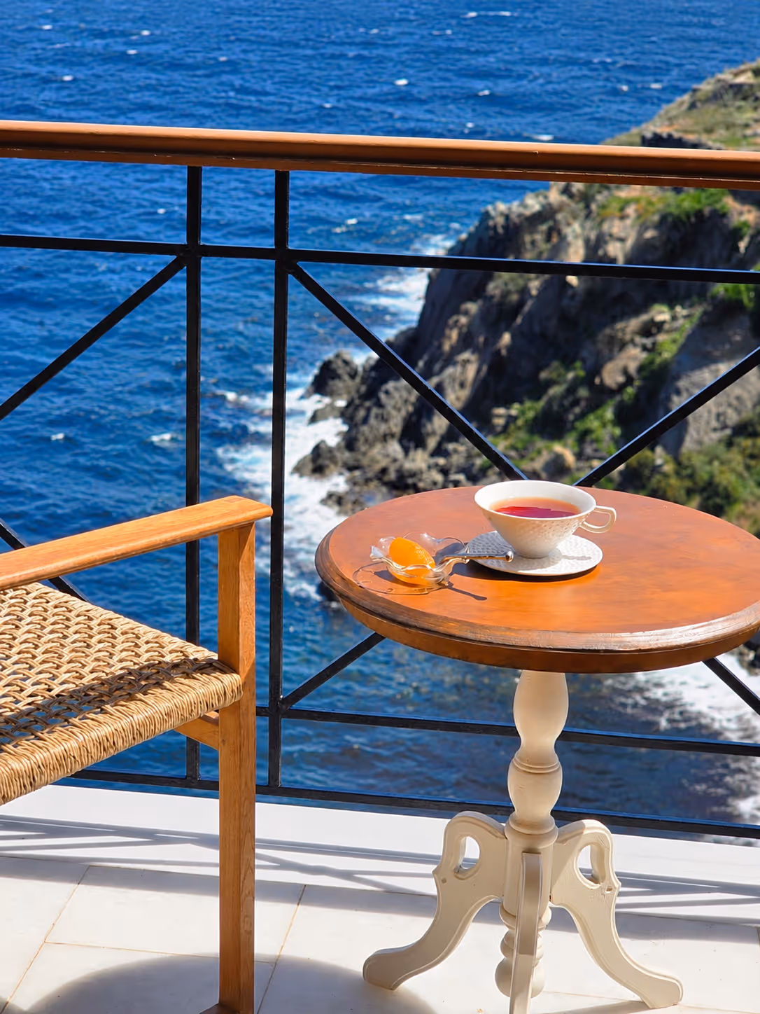 Wooden chair and round table with tea cup and orange slice overlooking rocky coastline and blue sea.