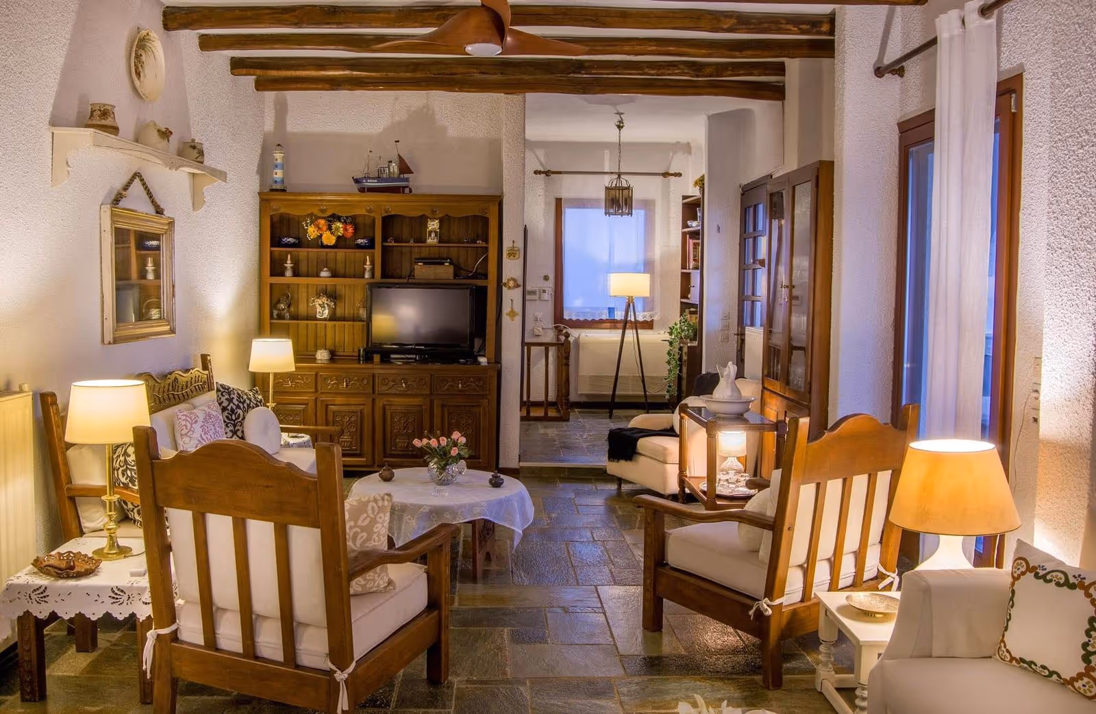 Cozy living room with warm lighting, wooden ceiling beams, cushioned wooden armchairs, a small dining table with a lace tablecloth, and a wooden cabinet with a TV.